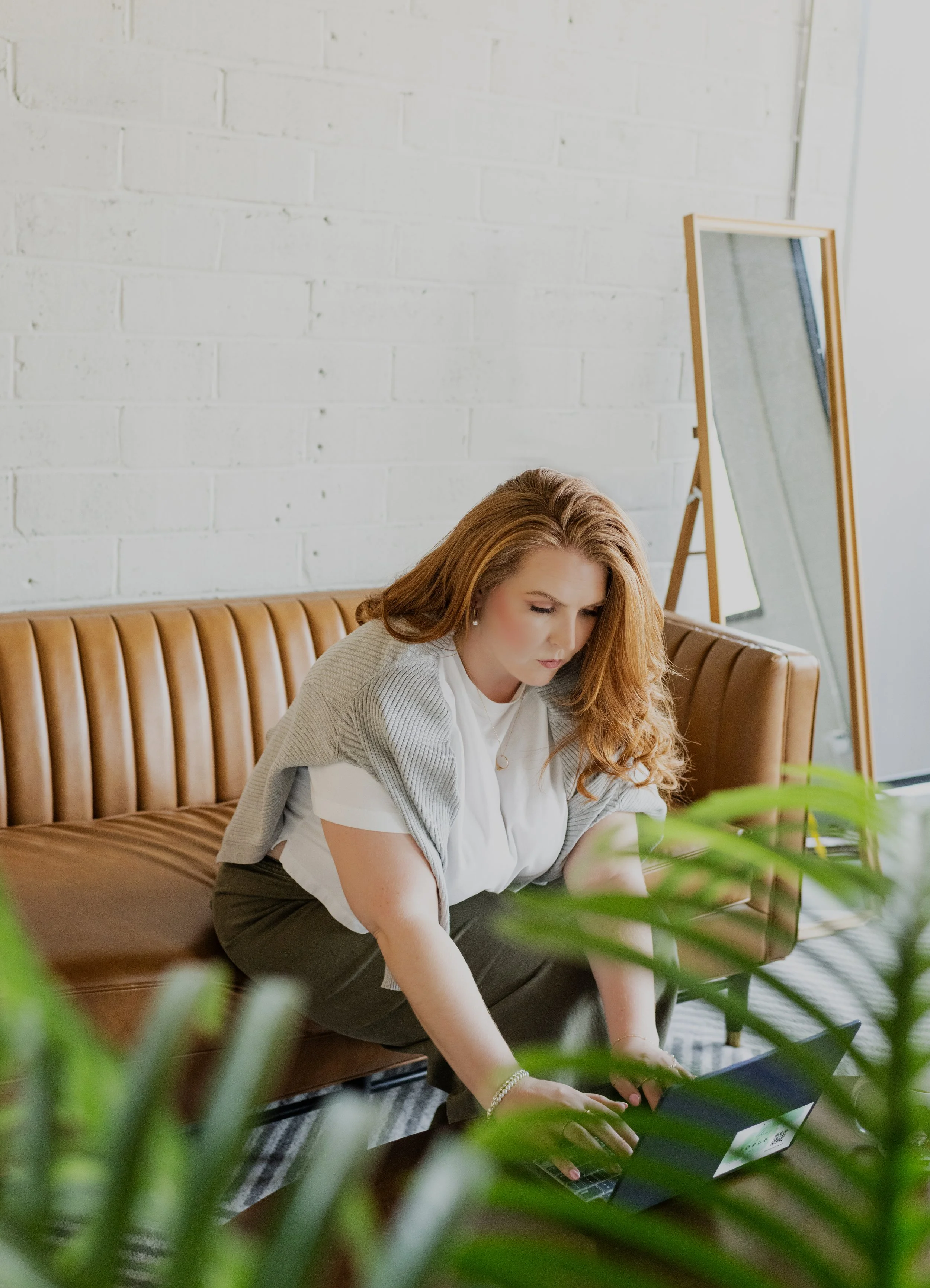 A woman with red hair sitting on a brown leather couch, working on a laptop, in a room with a white brick wall, a full-length mirror, and green plants.