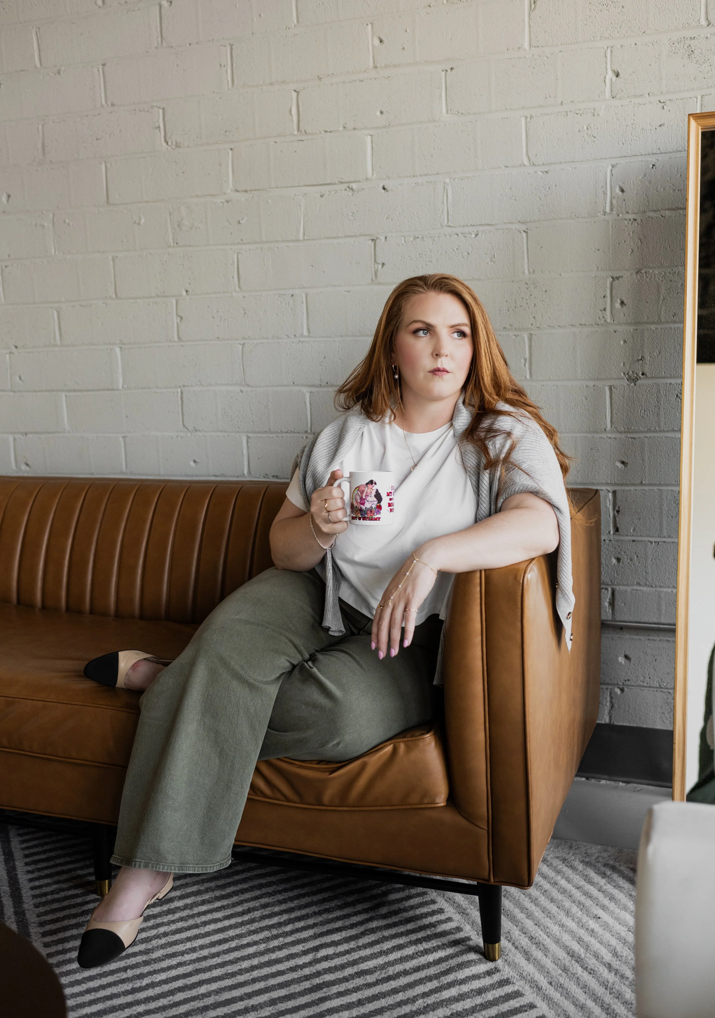 A woman with long red hair sitting on a brown leather sofa, holding a coffee mug, with a white brick wall behind her, looking to the right.