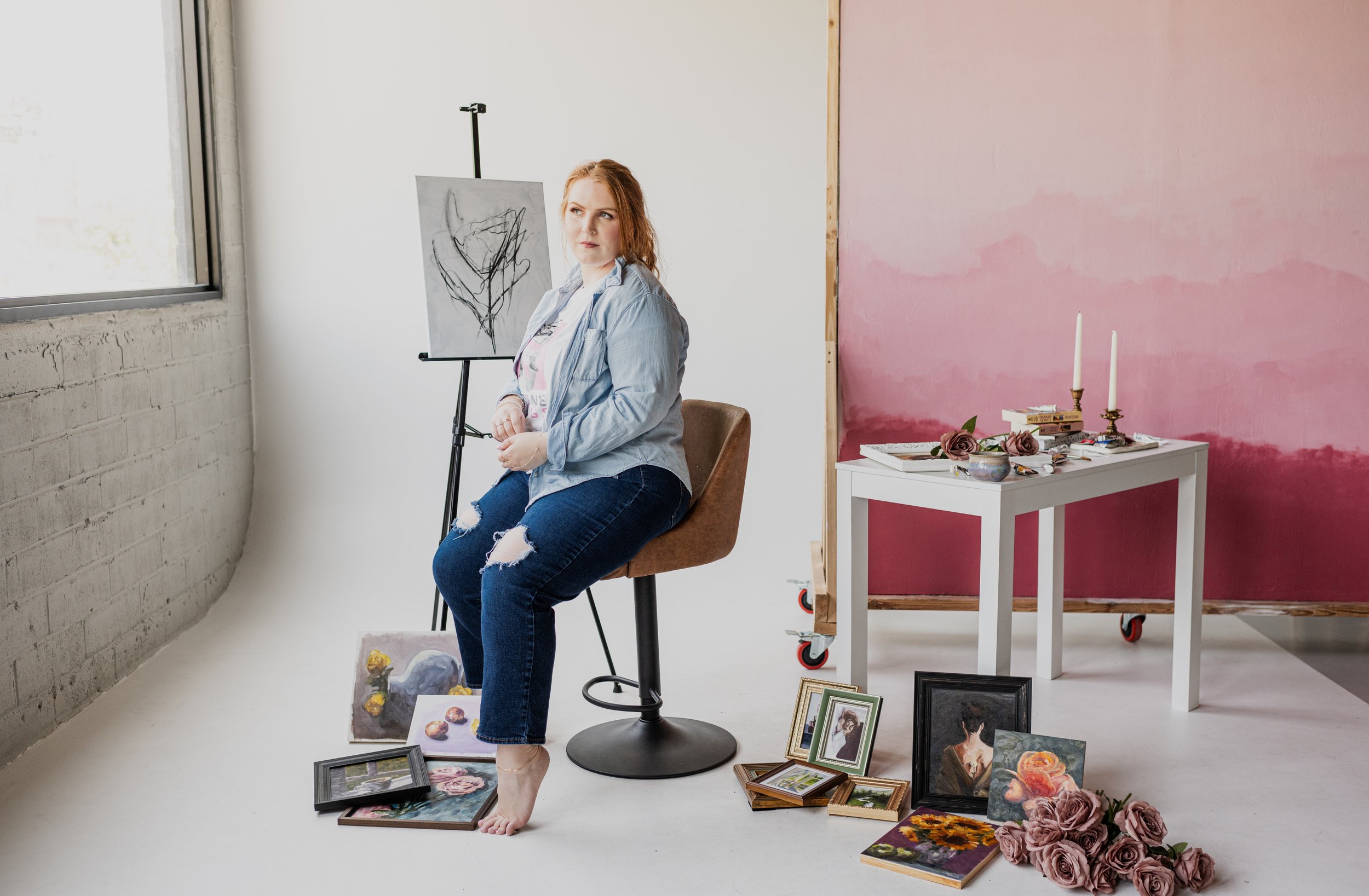Woman sitting on a chair in an art studio, surrounded by paintings, with a window to her left and an easel behind her. There are pictures on the floor and a table with candles, books, and flowers.