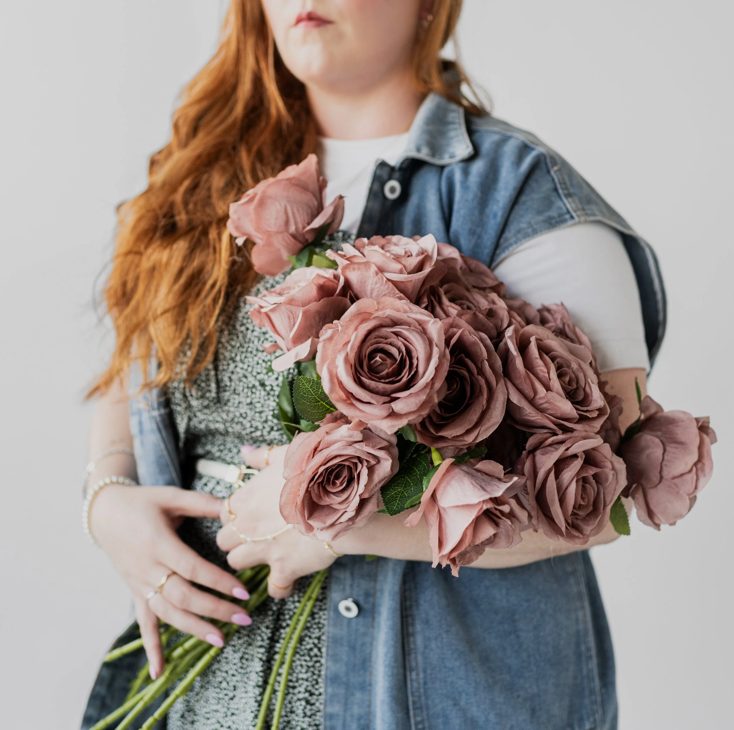 A woman with long red hair is holding a large bouquet of dusty pink roses and light pink peonies.
