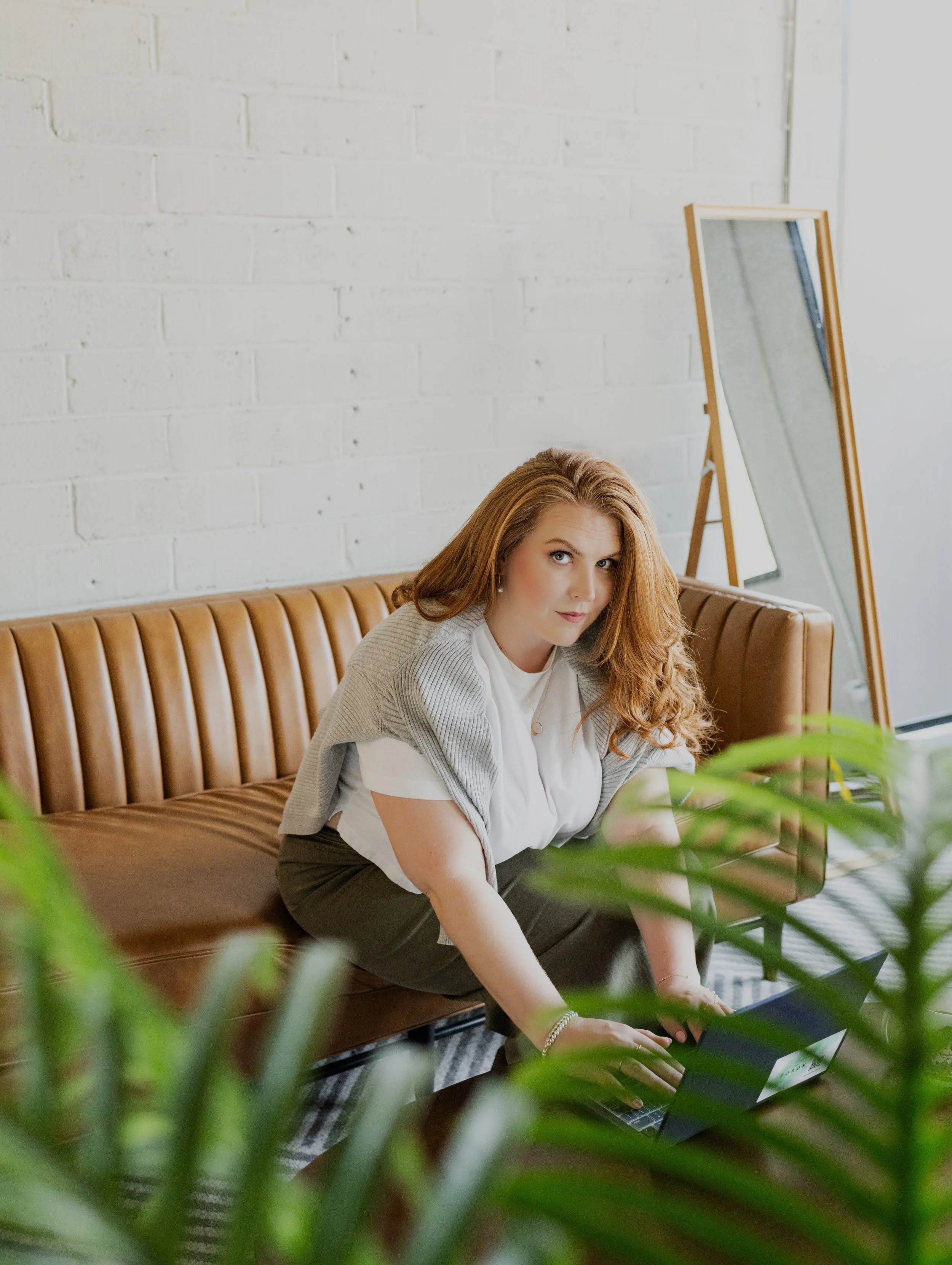 A woman with long, wavy red hair sitting on a tan leather couch, working on a laptop, in a room with a white brick wall and a freestanding mirror.