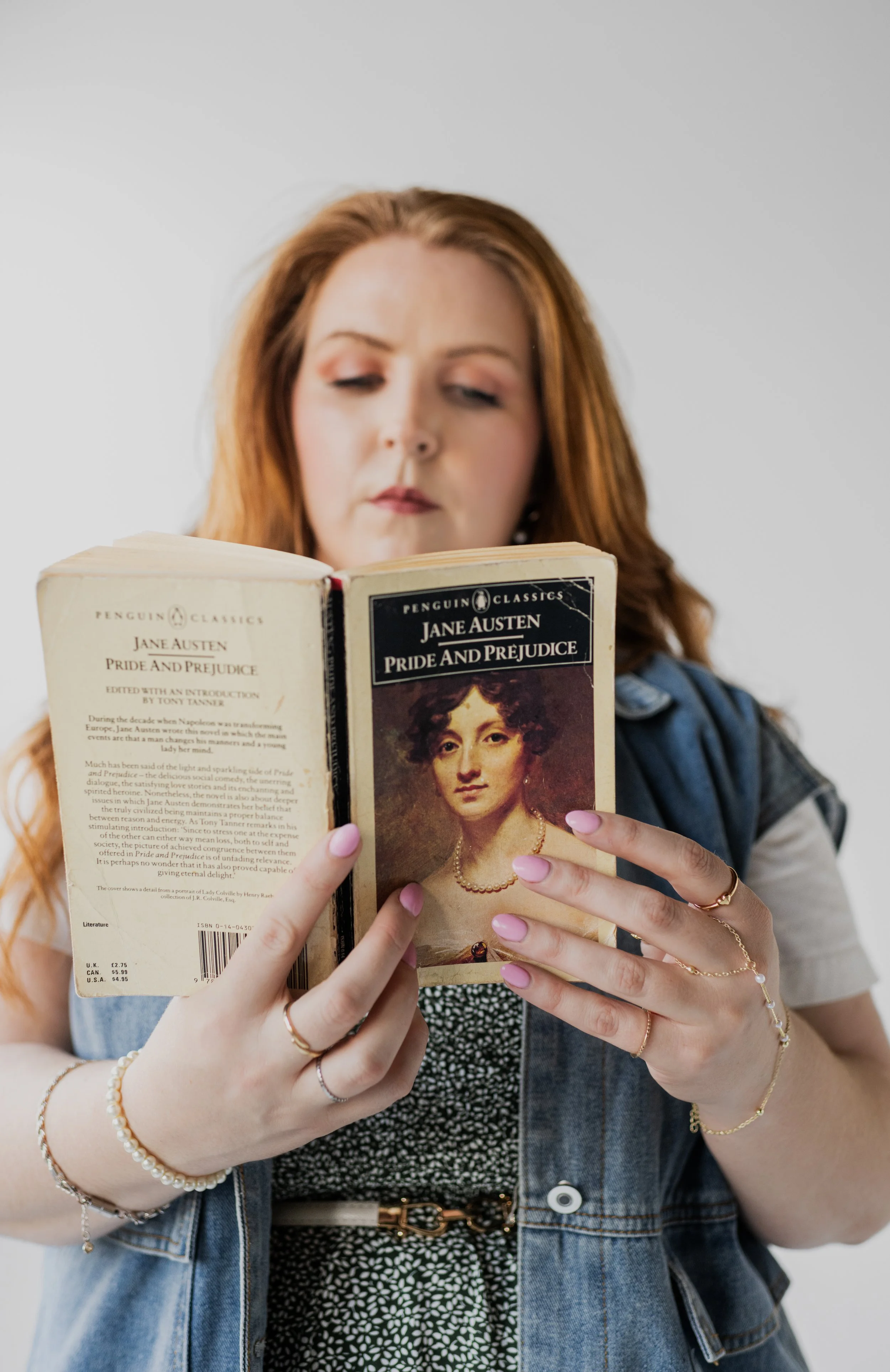 A woman with red hair holding a paperback copy of Jane Austen's Pride and Prejudice, focused on reading.