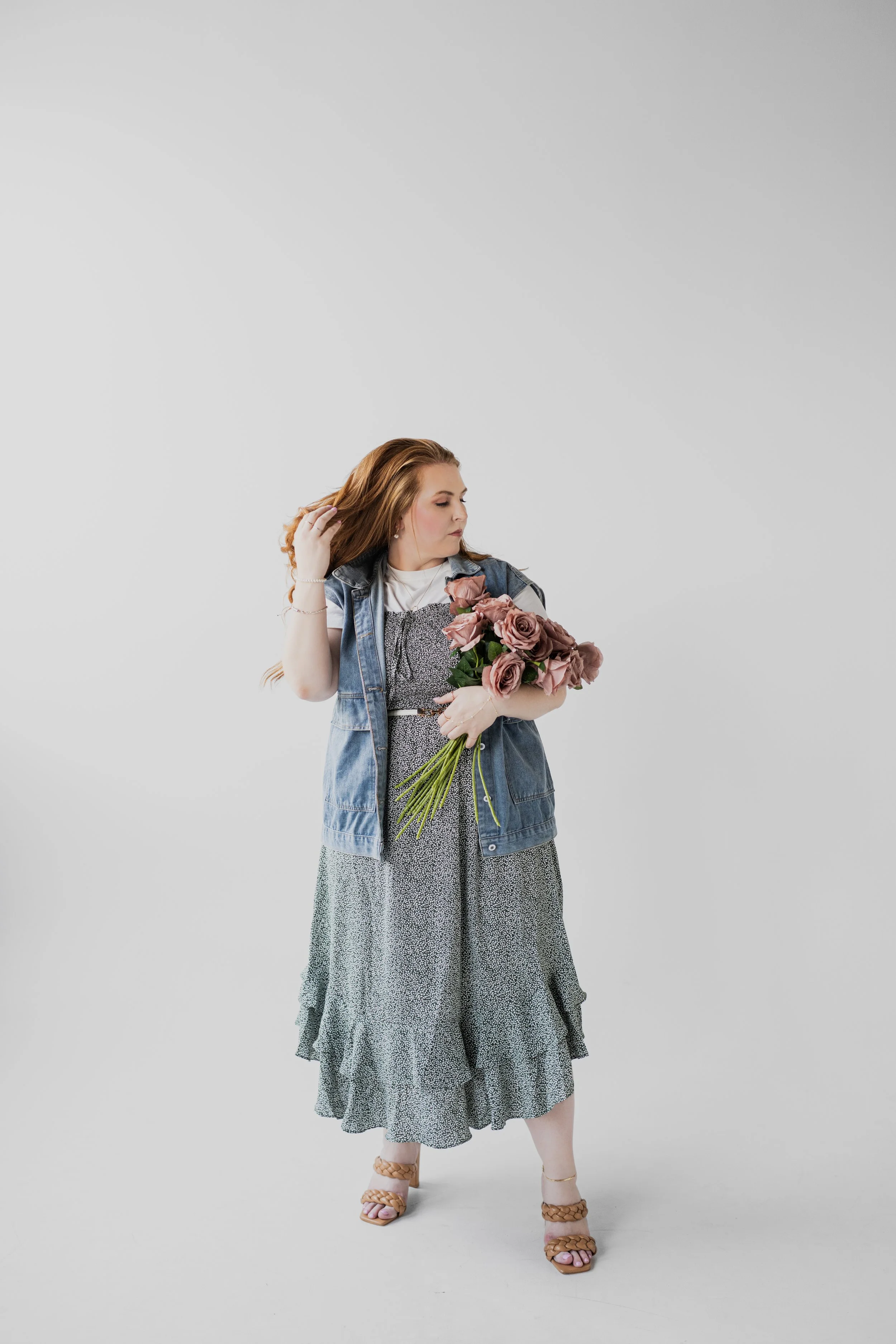 A woman with long red hair in a floral dress, denim vest, and sandals holding a bouquet of pink roses against a plain white background.