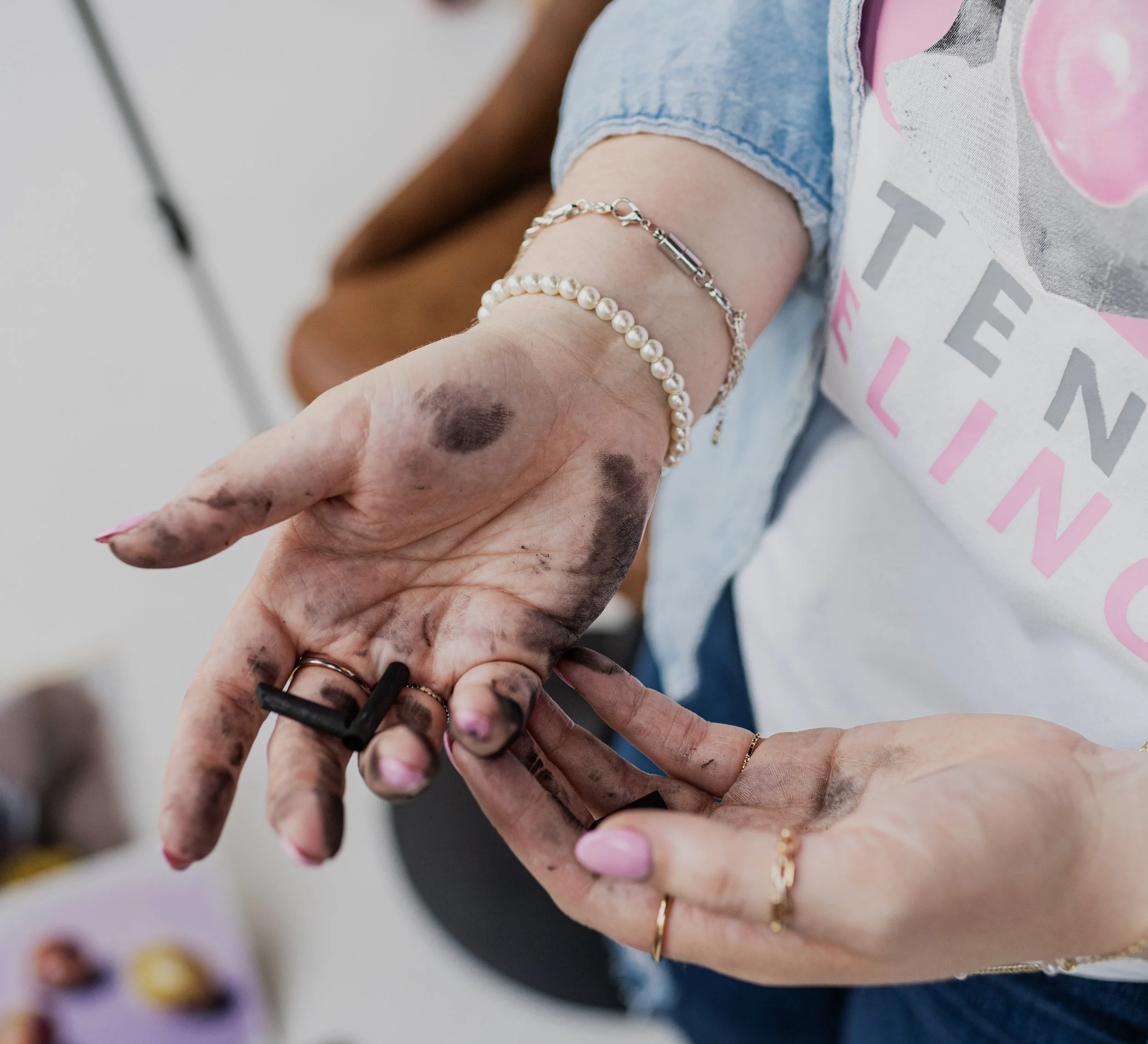 Close-up of a person's dirty and ink-stained hand with black ink on fingers, holding a small black marker, next to another hand with pink nail polish and jewelry, in an indoor setting.