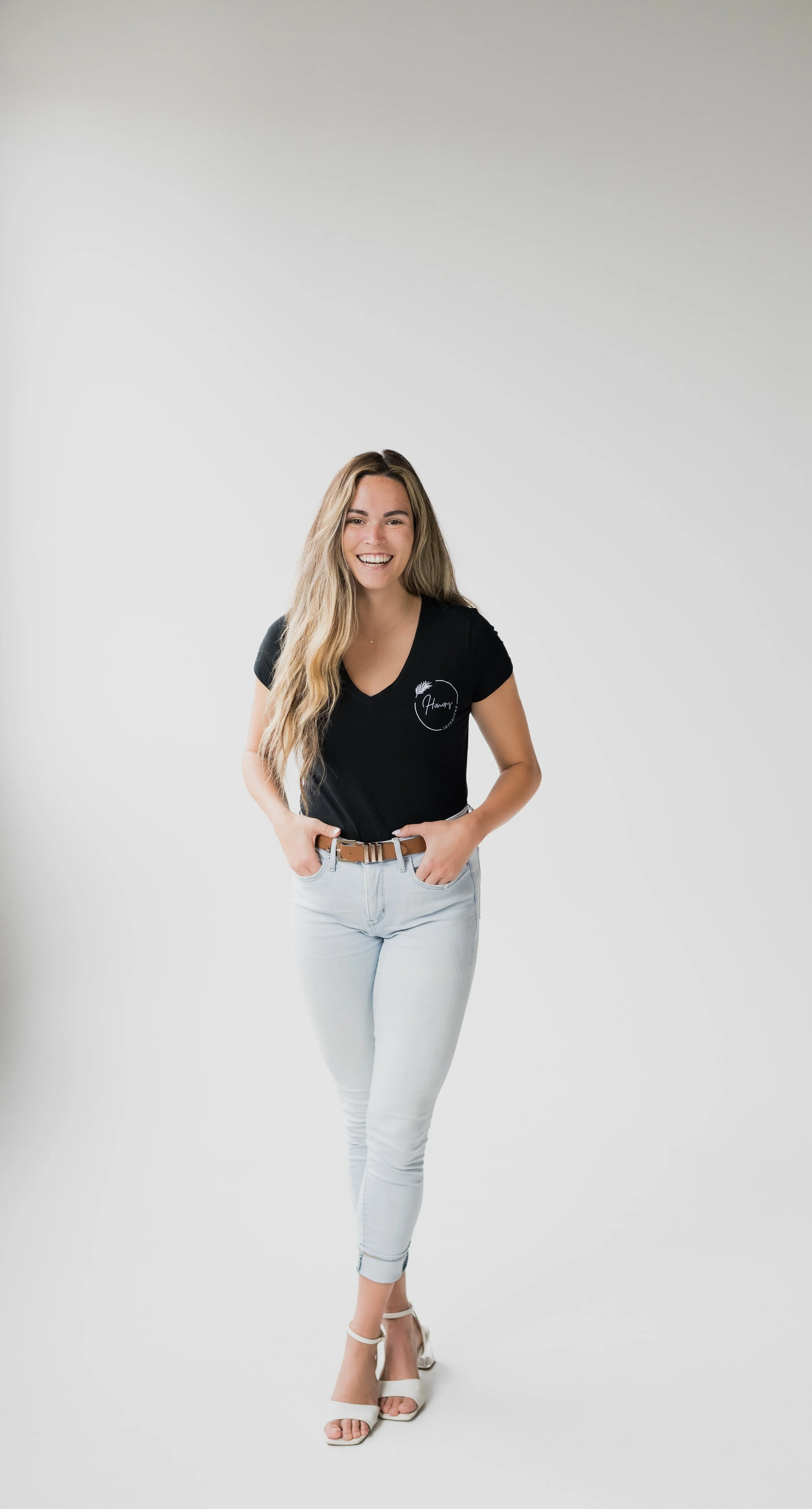 A young woman with long blonde hair, smiling and standing with her hands in her pockets against a plain white background, wearing a black T-shirt, light jeans, and white high-heeled sandals.