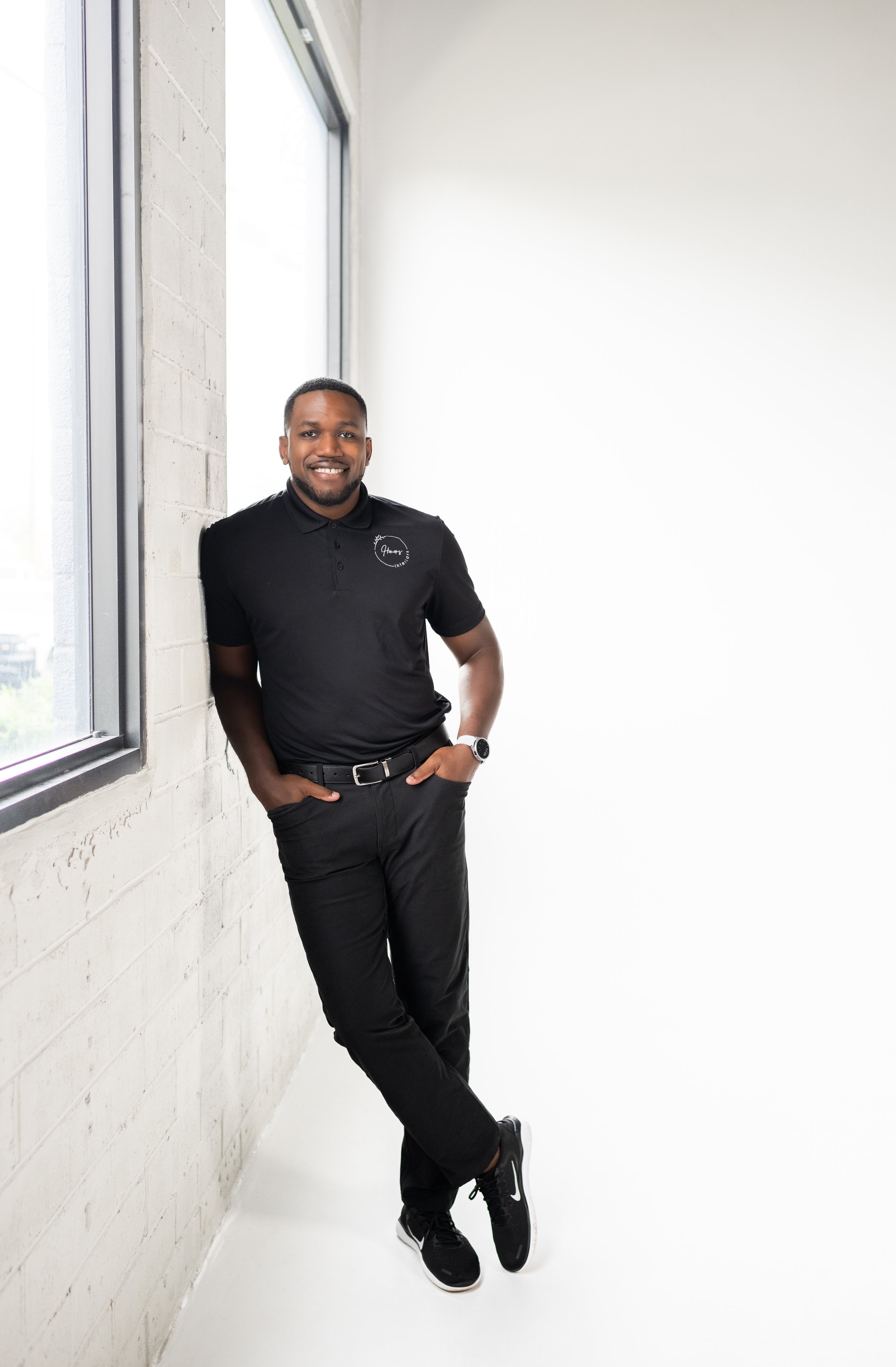 A smiling man in black polo shirt and black pants standing by a large window in a bright room.