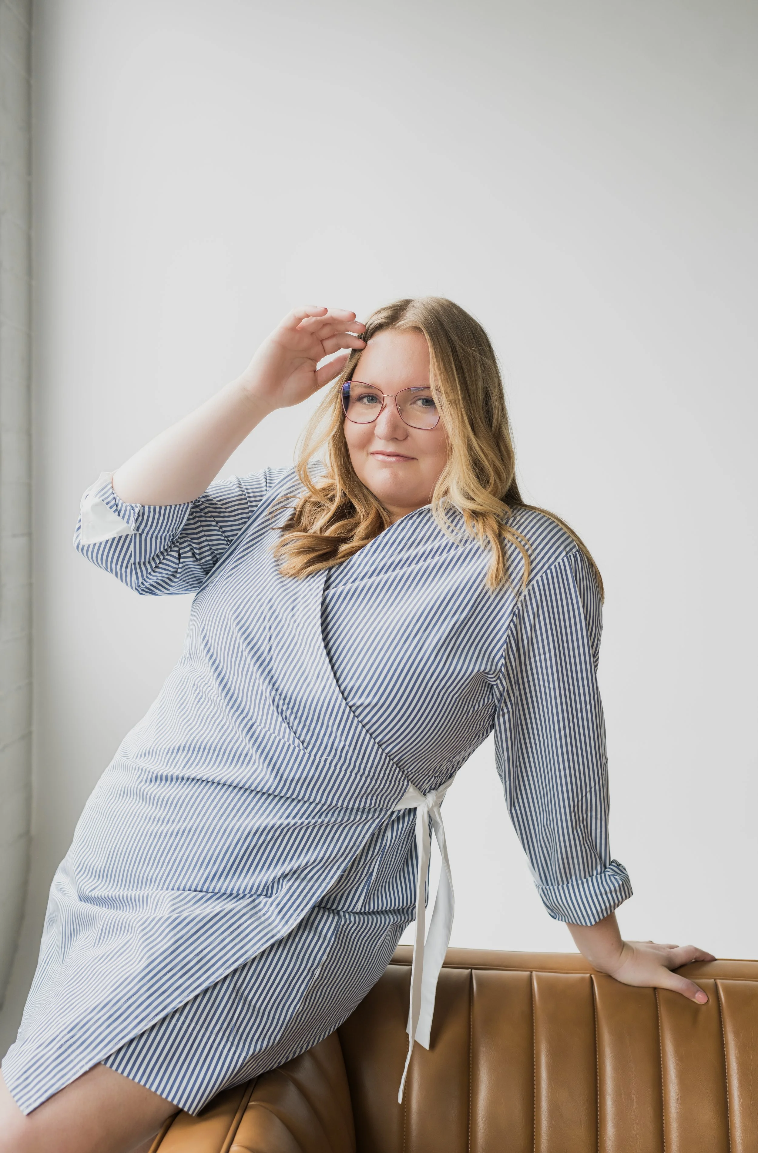 A woman with blonde hair wearing glasses and a blue and white striped dress, posed sitting on a brown leather bench and touching her head.