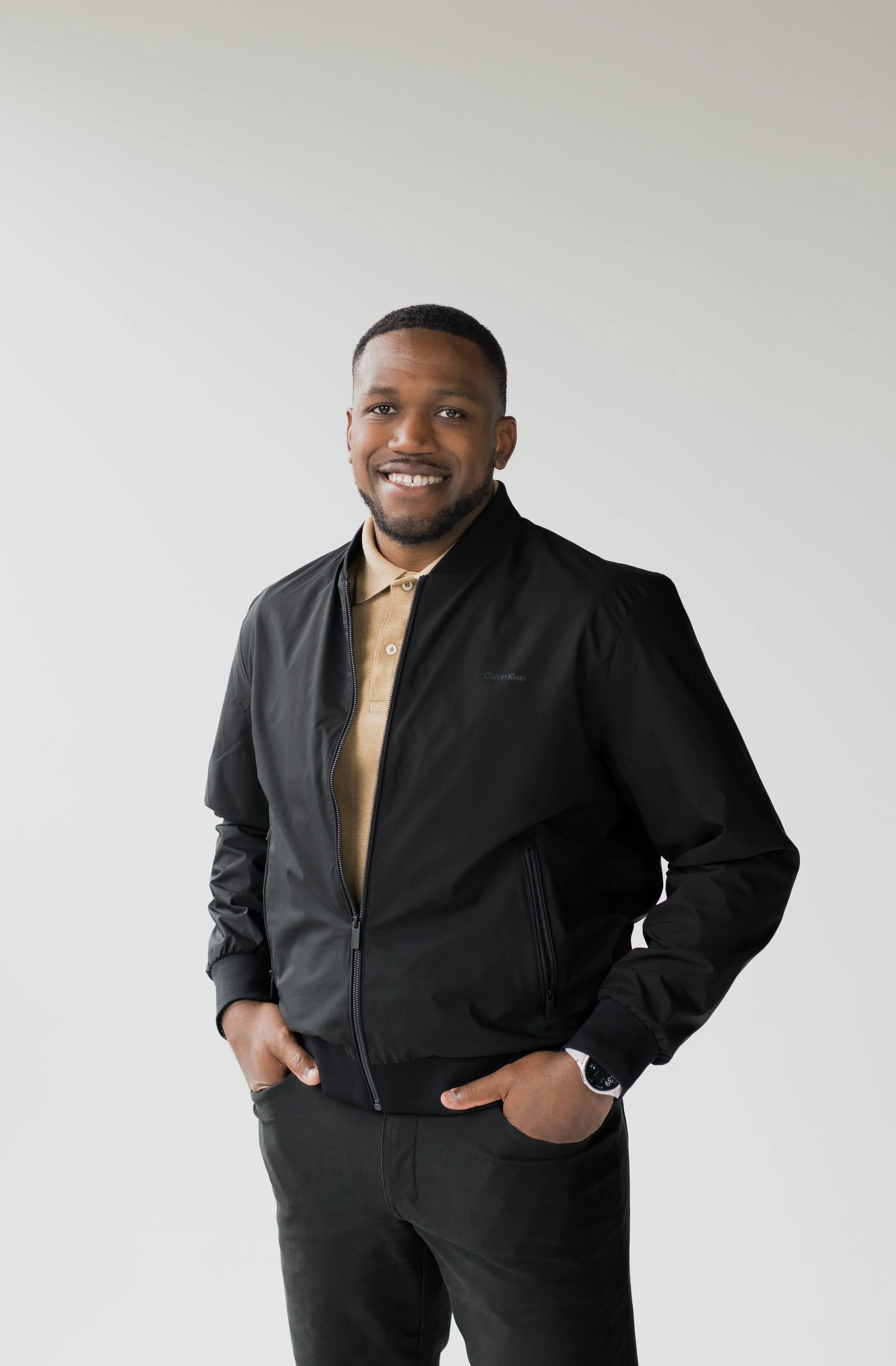 A smiling man in a beige shirt and black jacket standing with hands in pockets against a plain light background.
