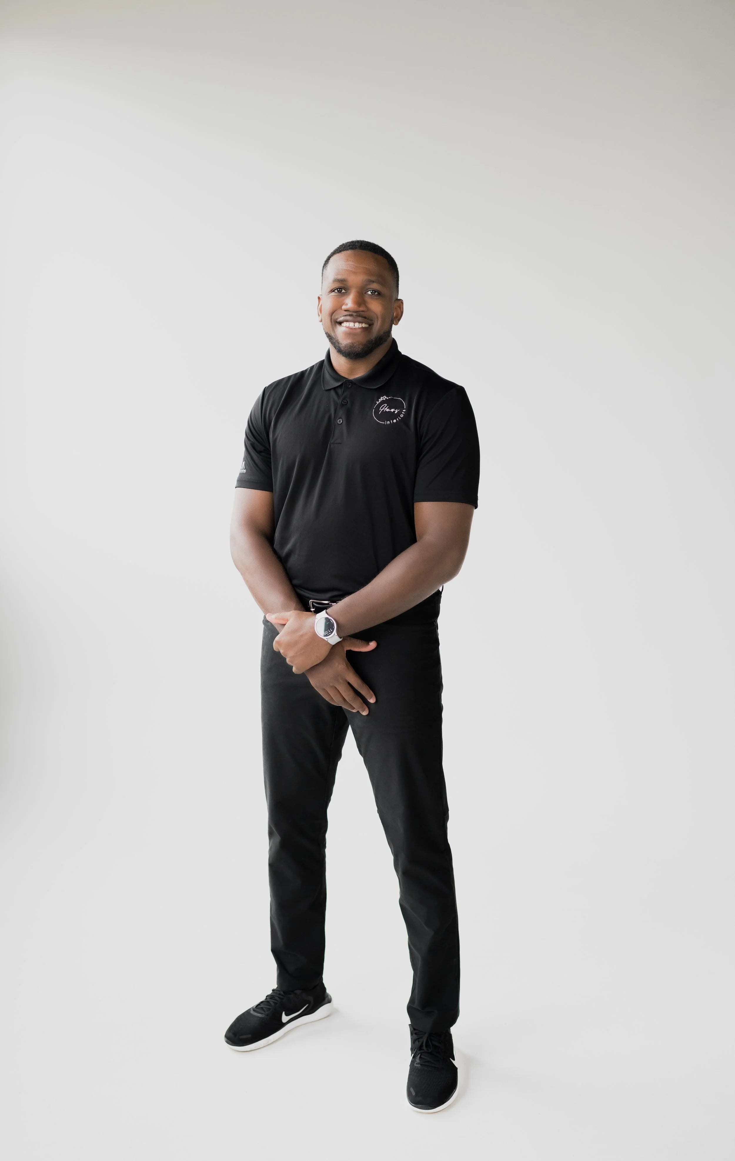 A smiling man in a black polo shirt, black pants, and black Nike sneakers standing against a plain white background.