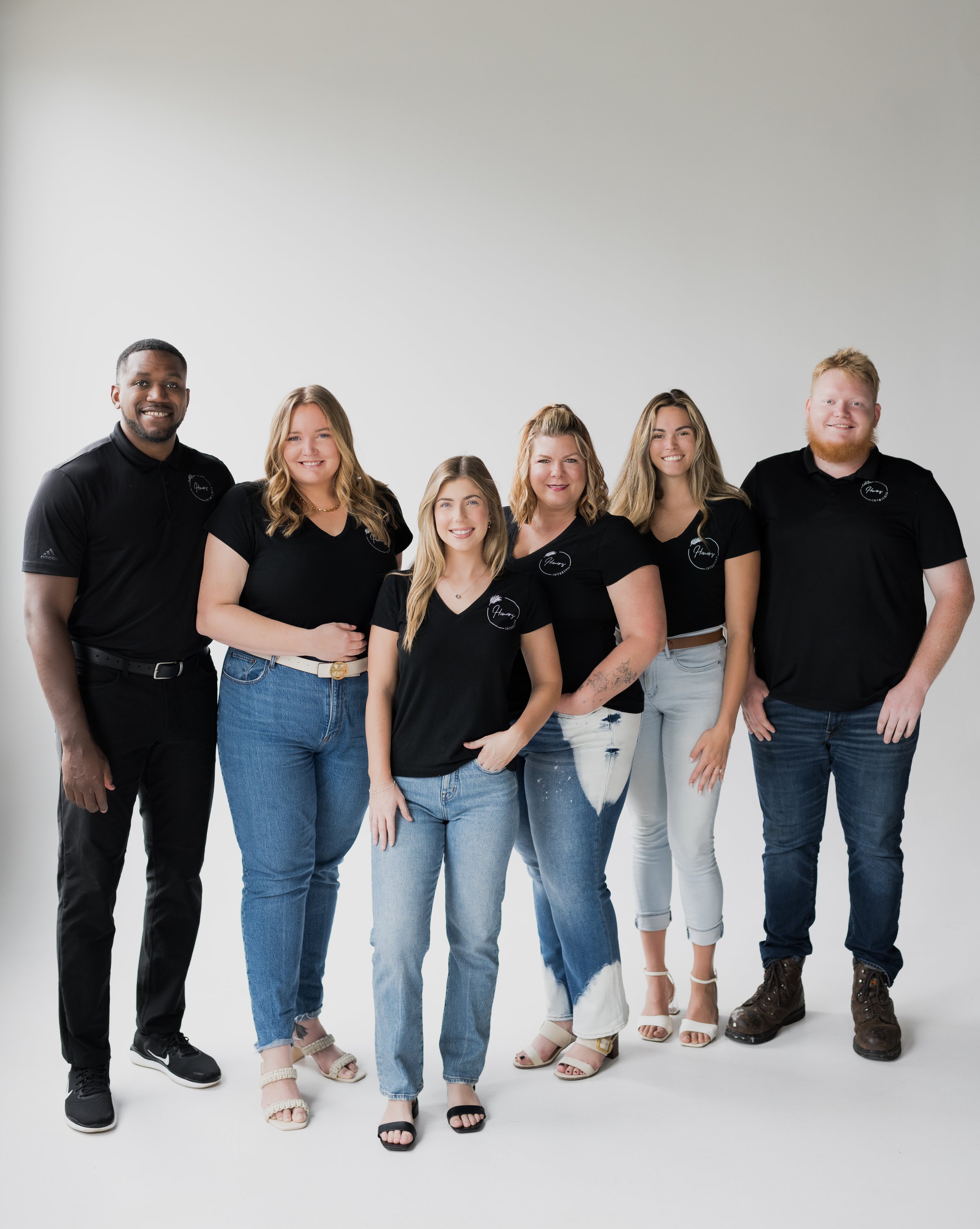Group of six people posing together in a photo studio with a white background, wearing black shirts with a logo.