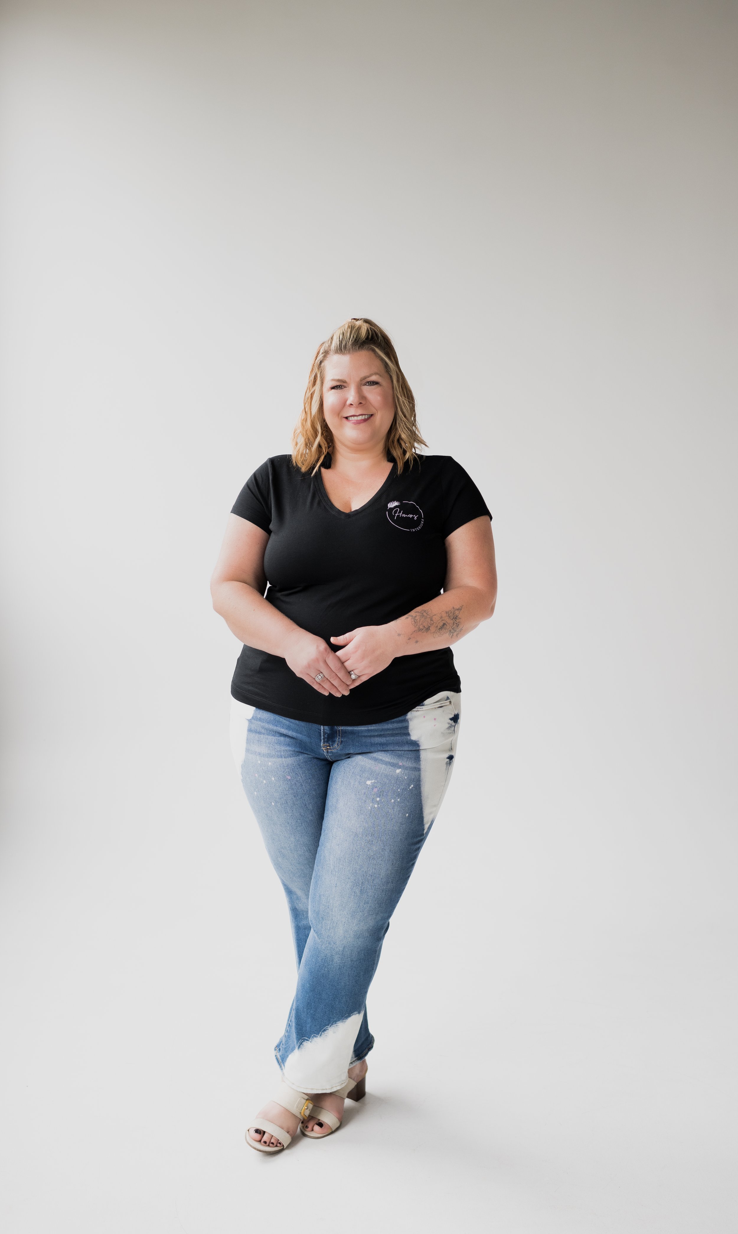 A woman with shoulder-length blond hair standing against a plain white background, wearing a black V-neck t-shirt, light blue distressed jeans with white paint splatters, and beige open-toe heels. She is smiling and has her hands clasped in front of 