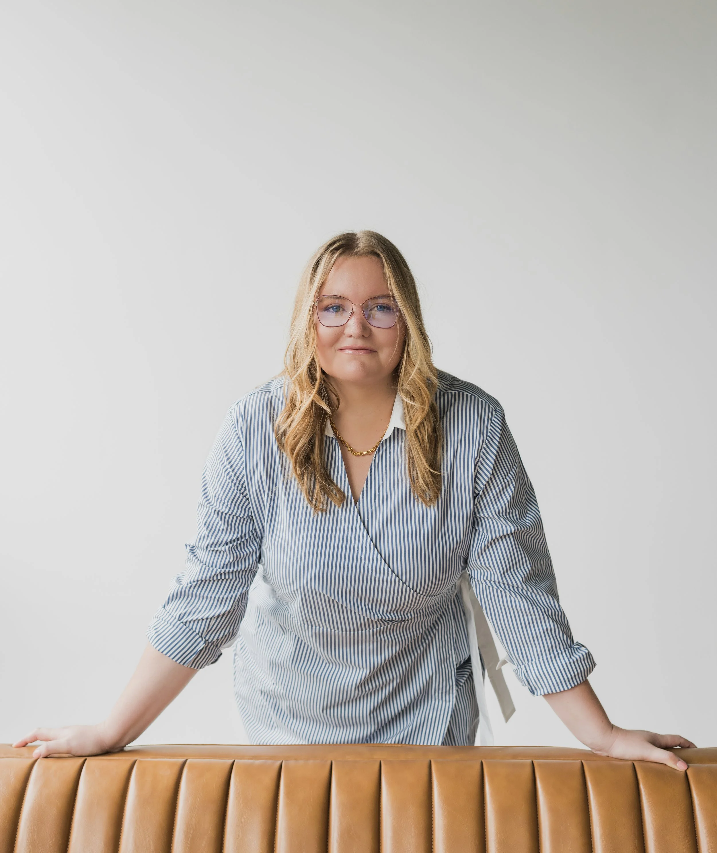 A woman with blonde hair, glasses, and a striped dress leaning on a tan leather bench against a plain white wall.