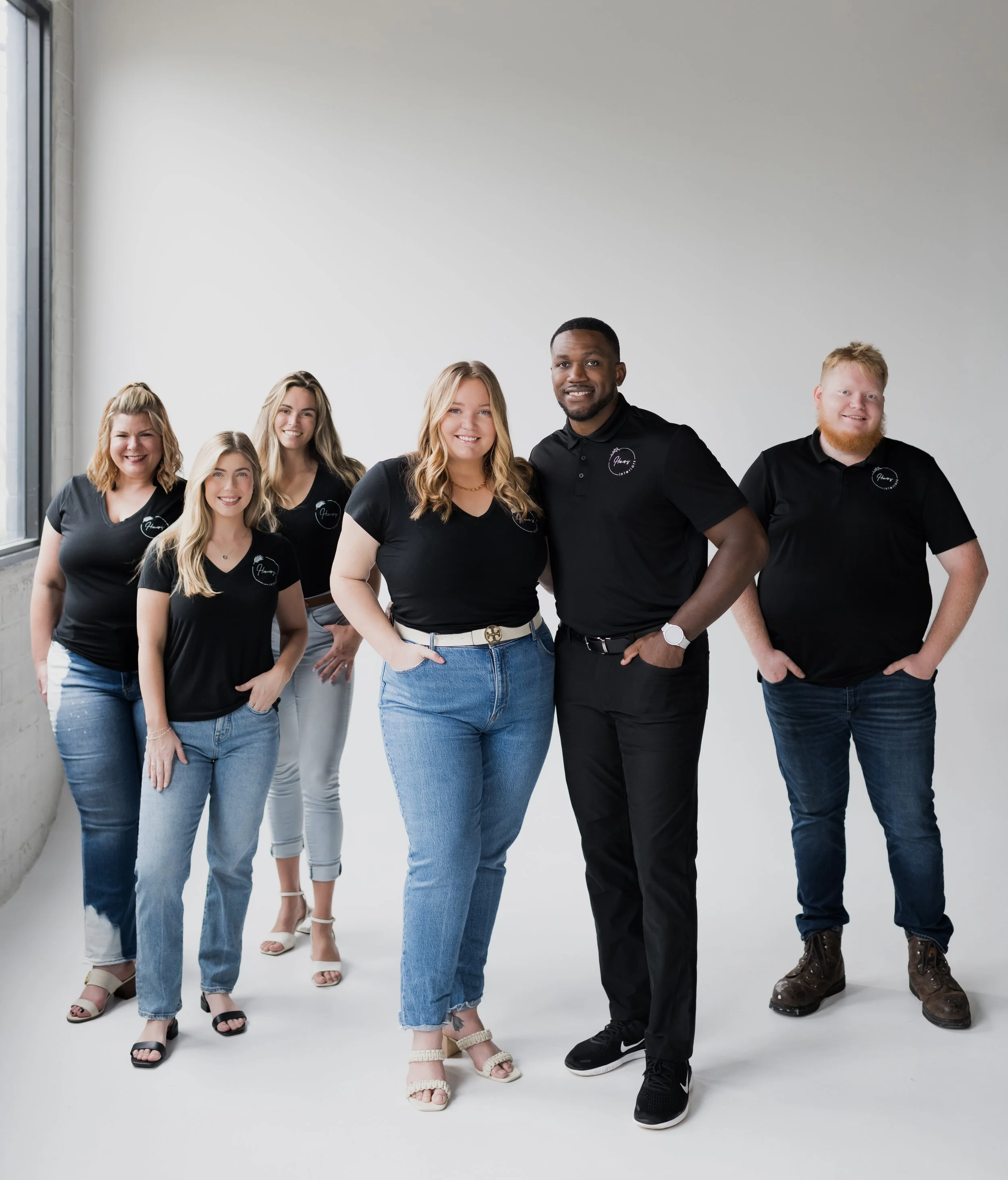 Group of six people standing together inside, wearing matching black shirts with logos, smiling for the photo.