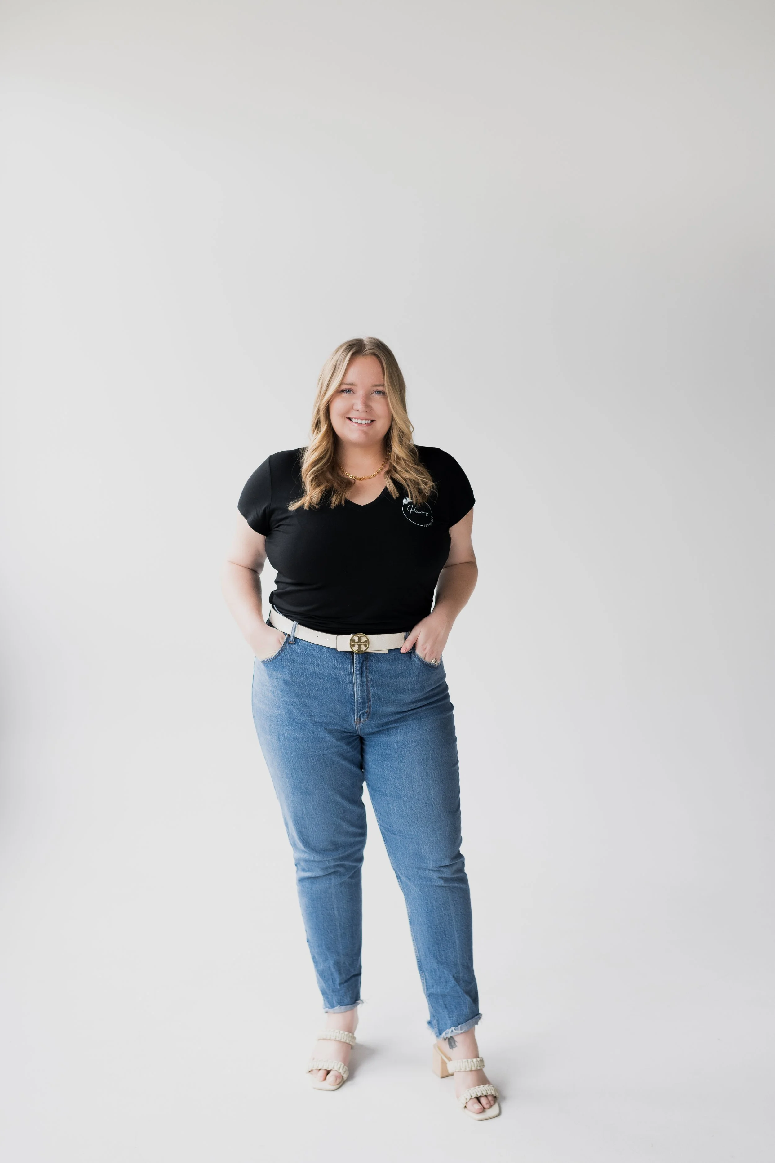 A woman with shoulder-length blonde hair wearing a black T-shirt, light blue jeans, white belt, and beige heeled sandals, standing against a plain white background, smiling at the camera.