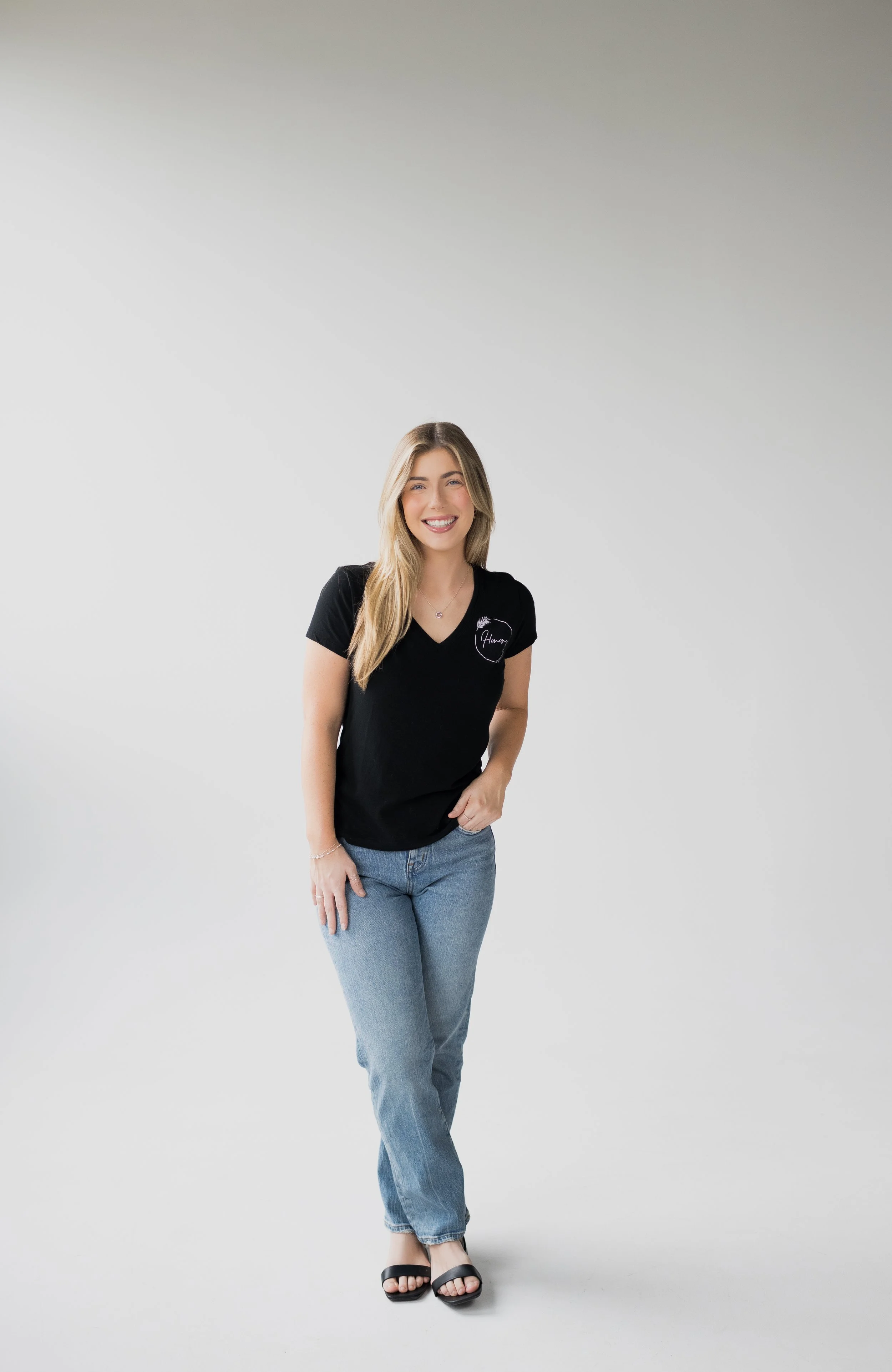 A young woman with long blonde hair, wearing a black T-shirt with a small logo, blue jeans, and black sandals, smiling and standing in front of a plain white background.