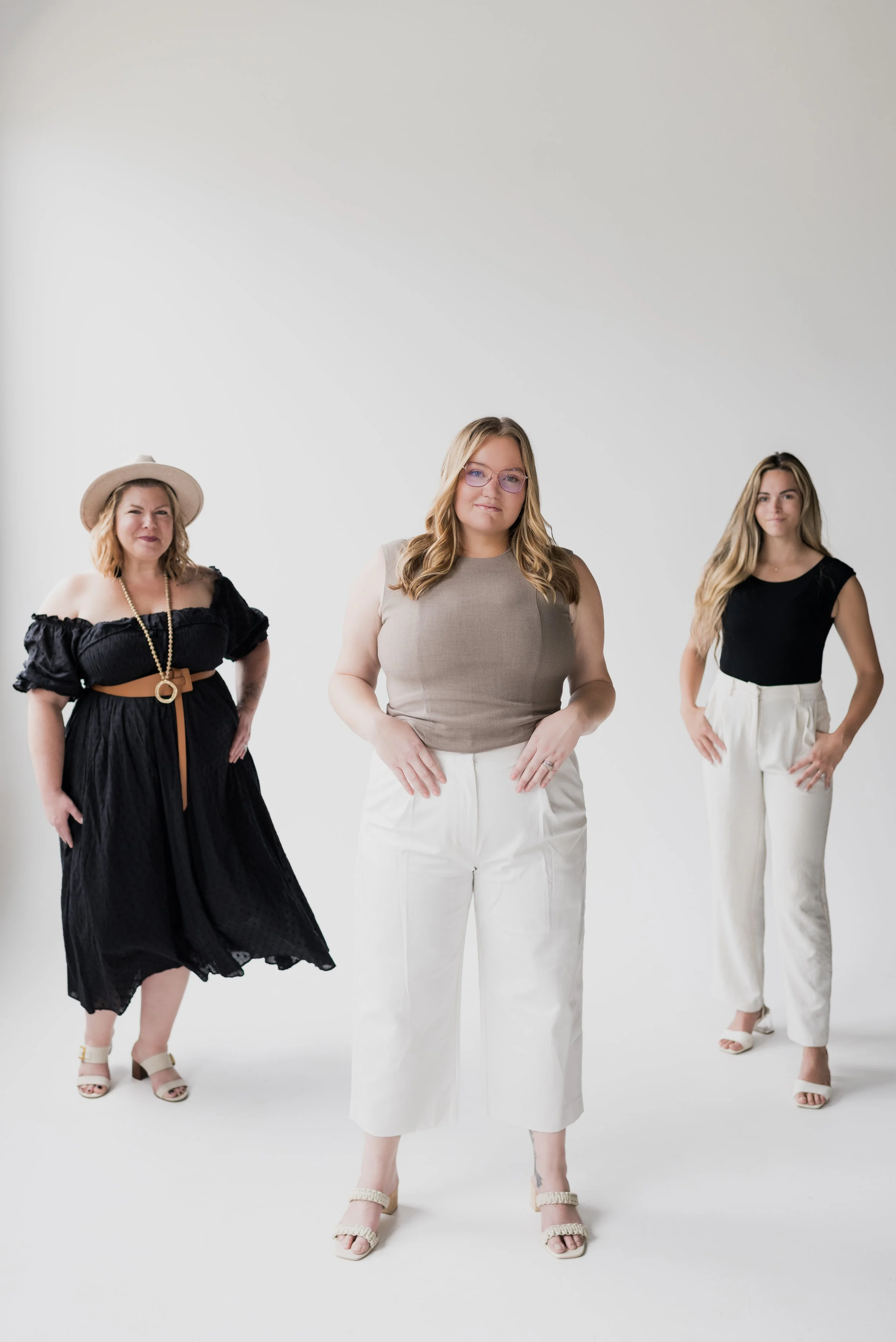 Three women standing against a plain white background, wearing stylish clothing and posing for the camera.