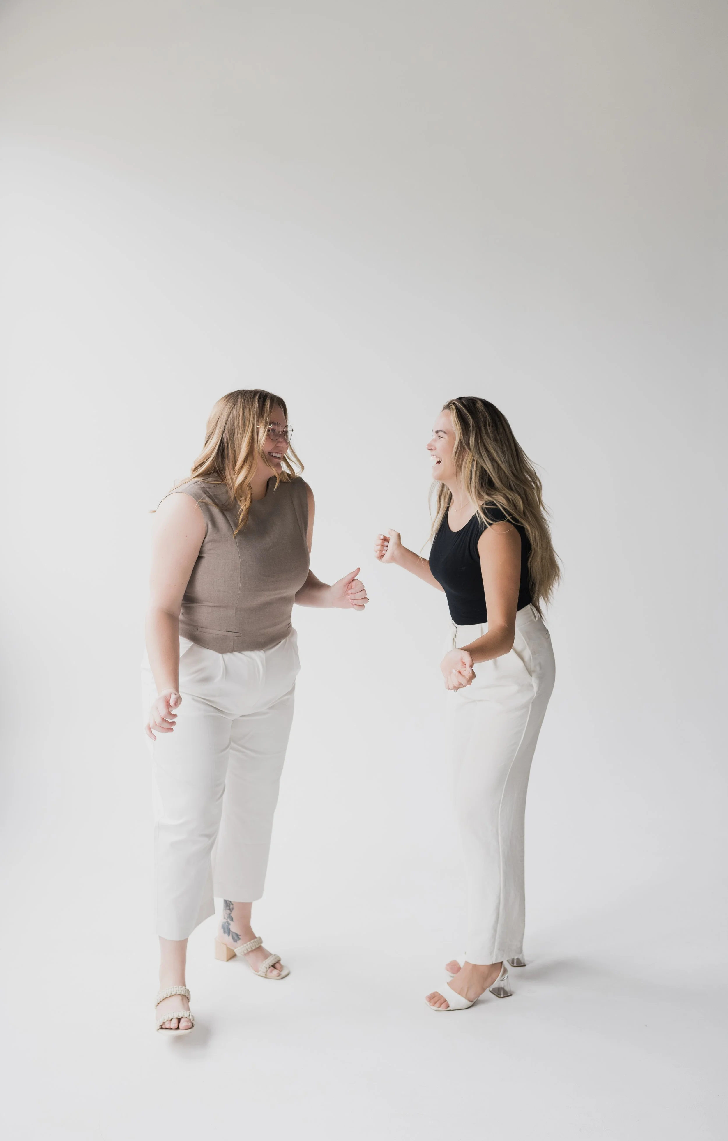 Two women standing facing each other, smiling and playingfully pretending to fight, on a plain white background.