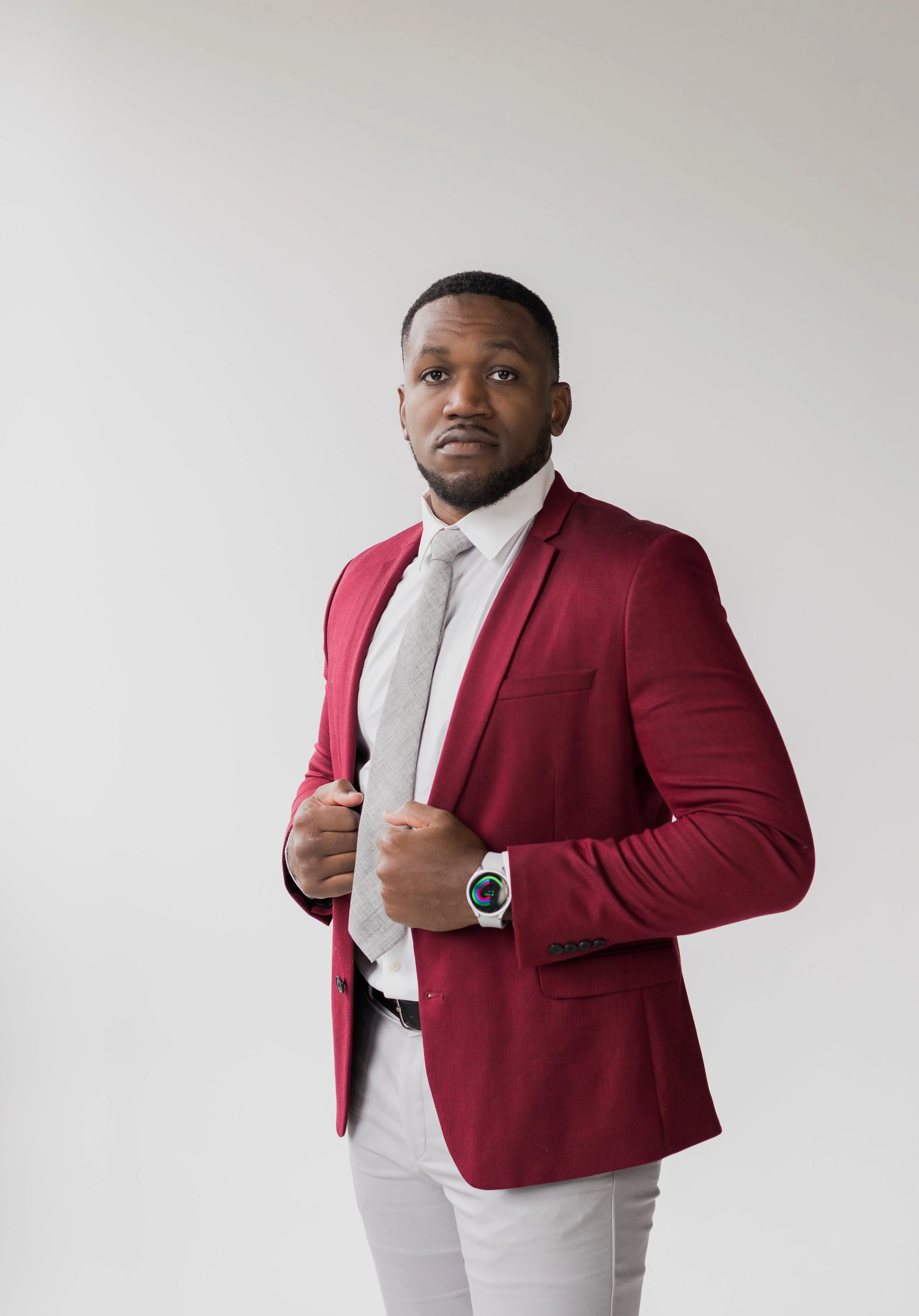 A man in a red blazer, white shirt, and light gray tie standing against a plain white background, holding the lapels of his blazer with both hands, wearing a smartwatch on his left wrist.