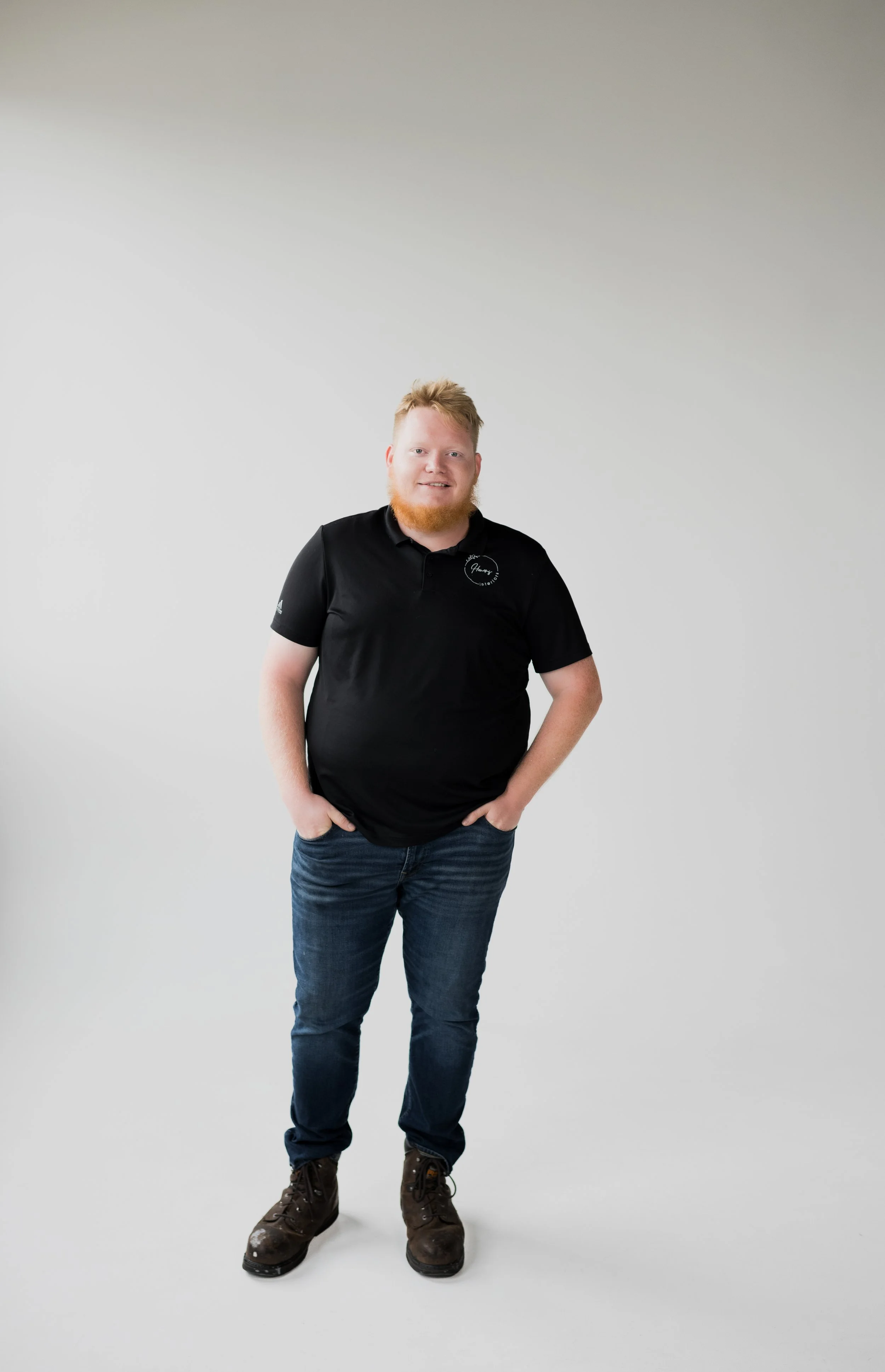 A man with red hair and beard standing against a plain white wall, wearing a black polo shirt, jeans, and brown work boots, with hands in his pockets and smiling at the camera.