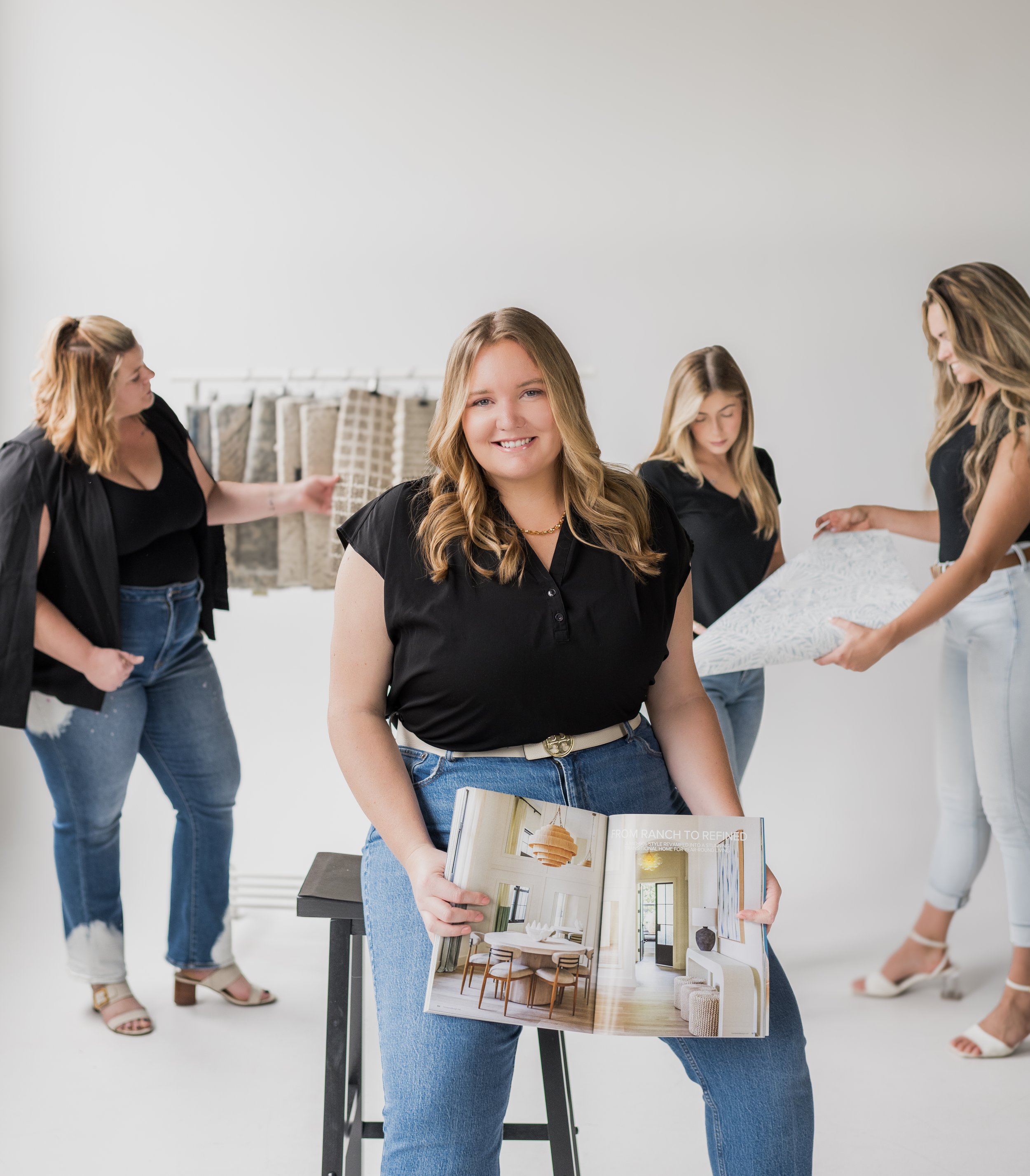 Young woman smiling while holding an interior design magazine, with four other women discussing fabric swatches and design plans in the background.