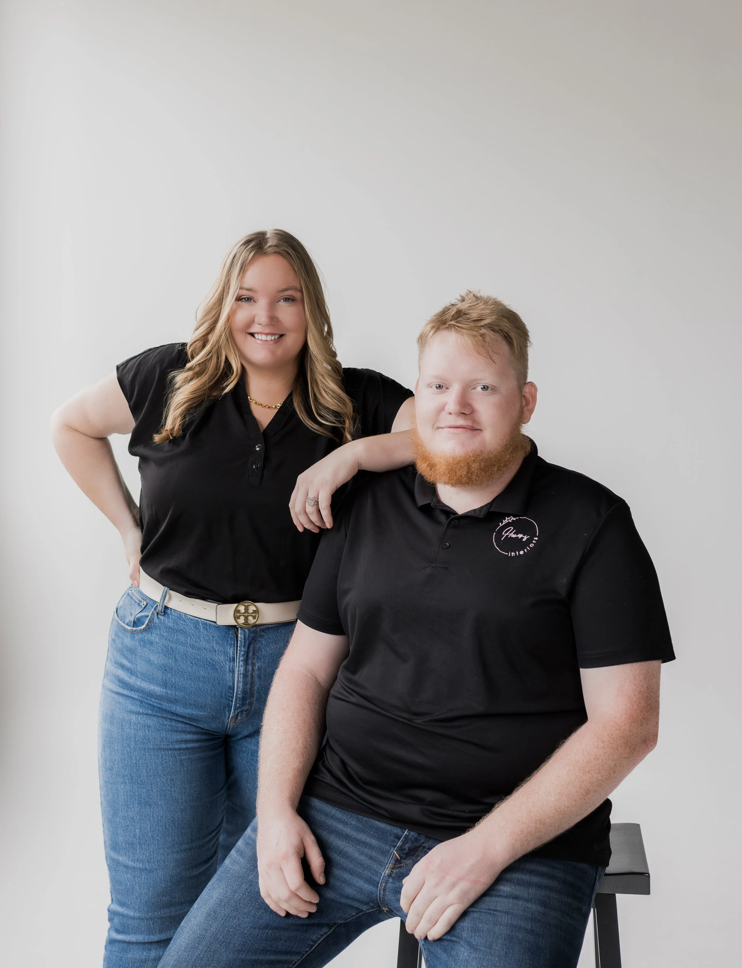 A smiling woman with long wavy hair and a man with a beard pose together against a plain light gray background. The woman has her arm resting on the man's shoulder, and they are both wearing black shirts and blue jeans.