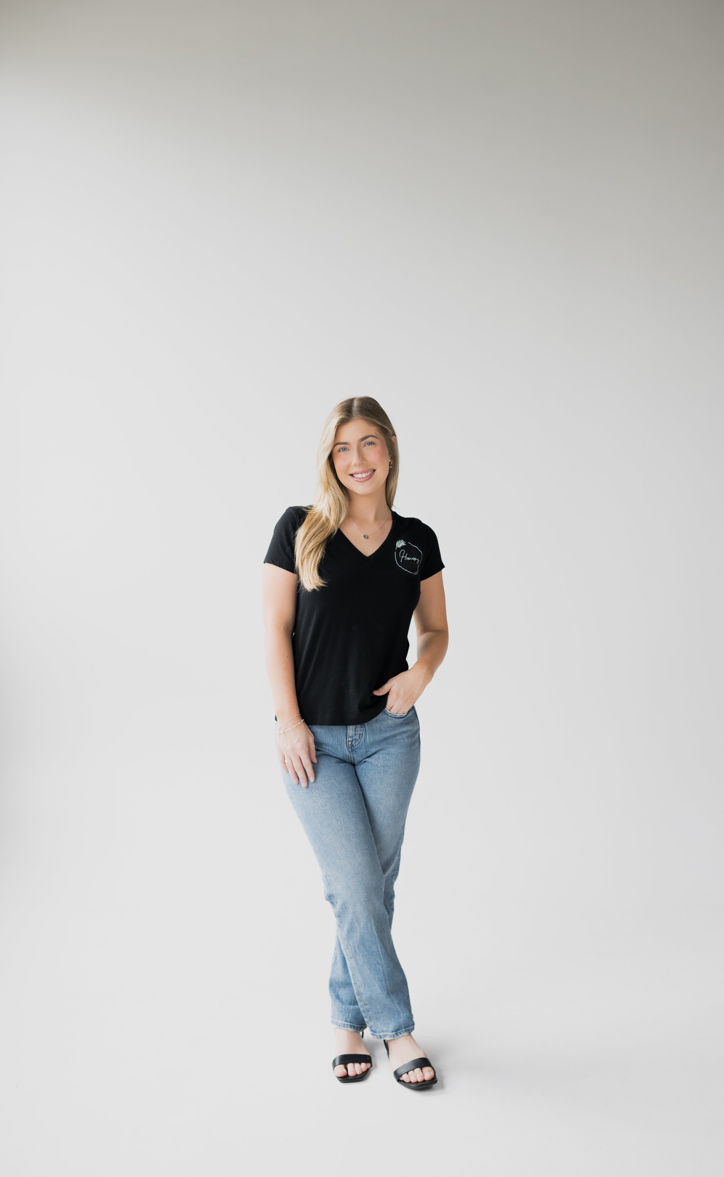 Young woman with long blonde hair wearing a black t-shirt and jeans, standing and smiling against a plain background.