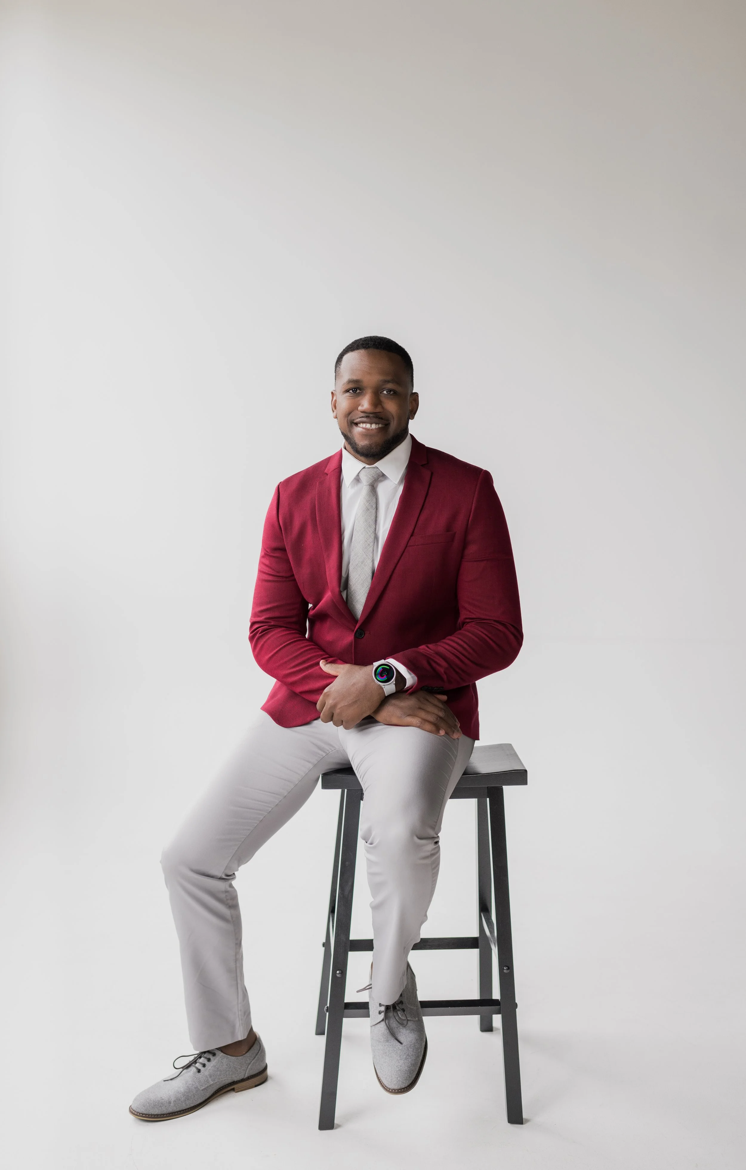 A man in a red blazer, white shirt, and grey pants sitting on a stool against a white background, smiling at the camera.