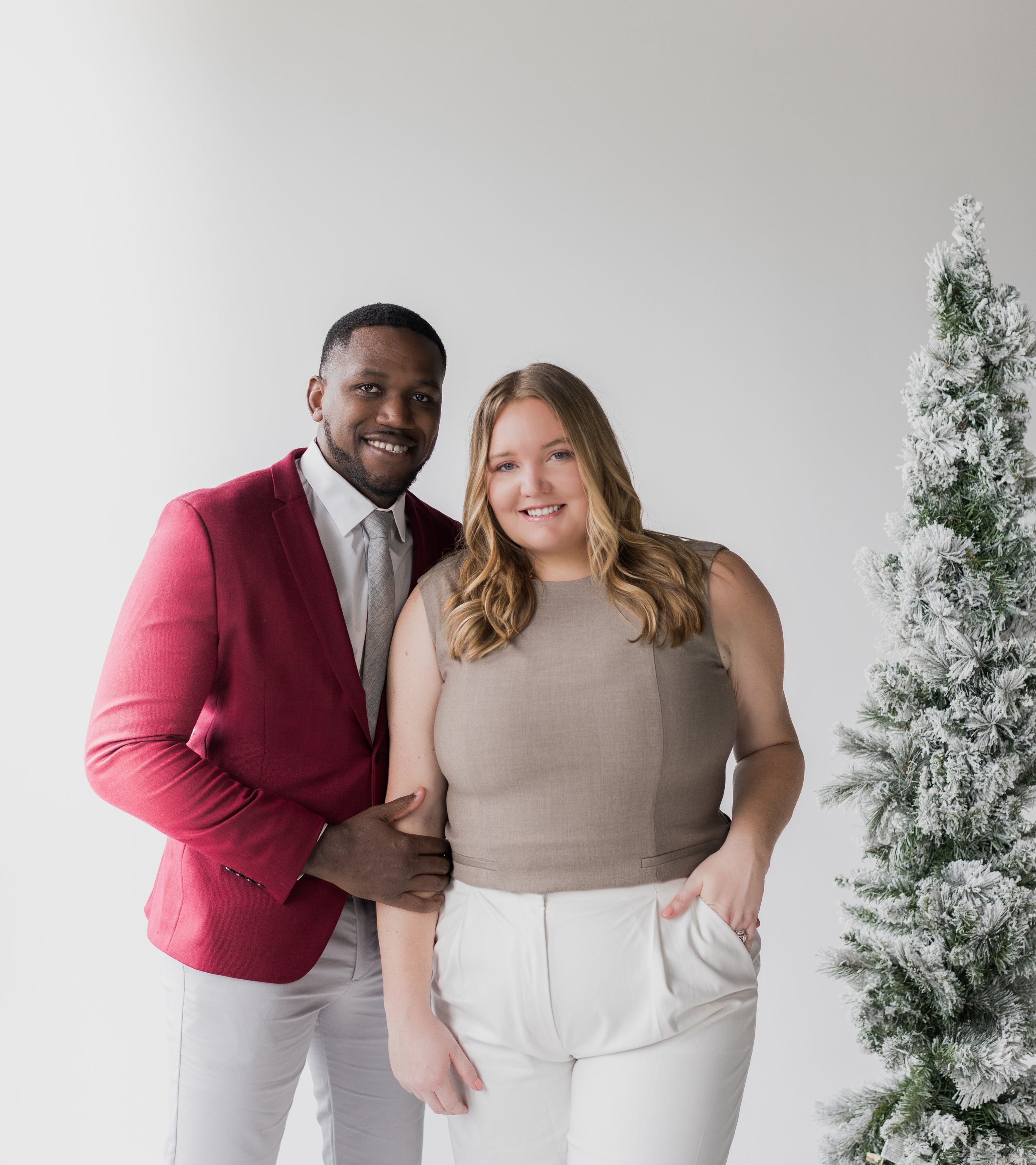A man and a woman standing side by side, smiling, with a snow-covered Christmas tree in the background.