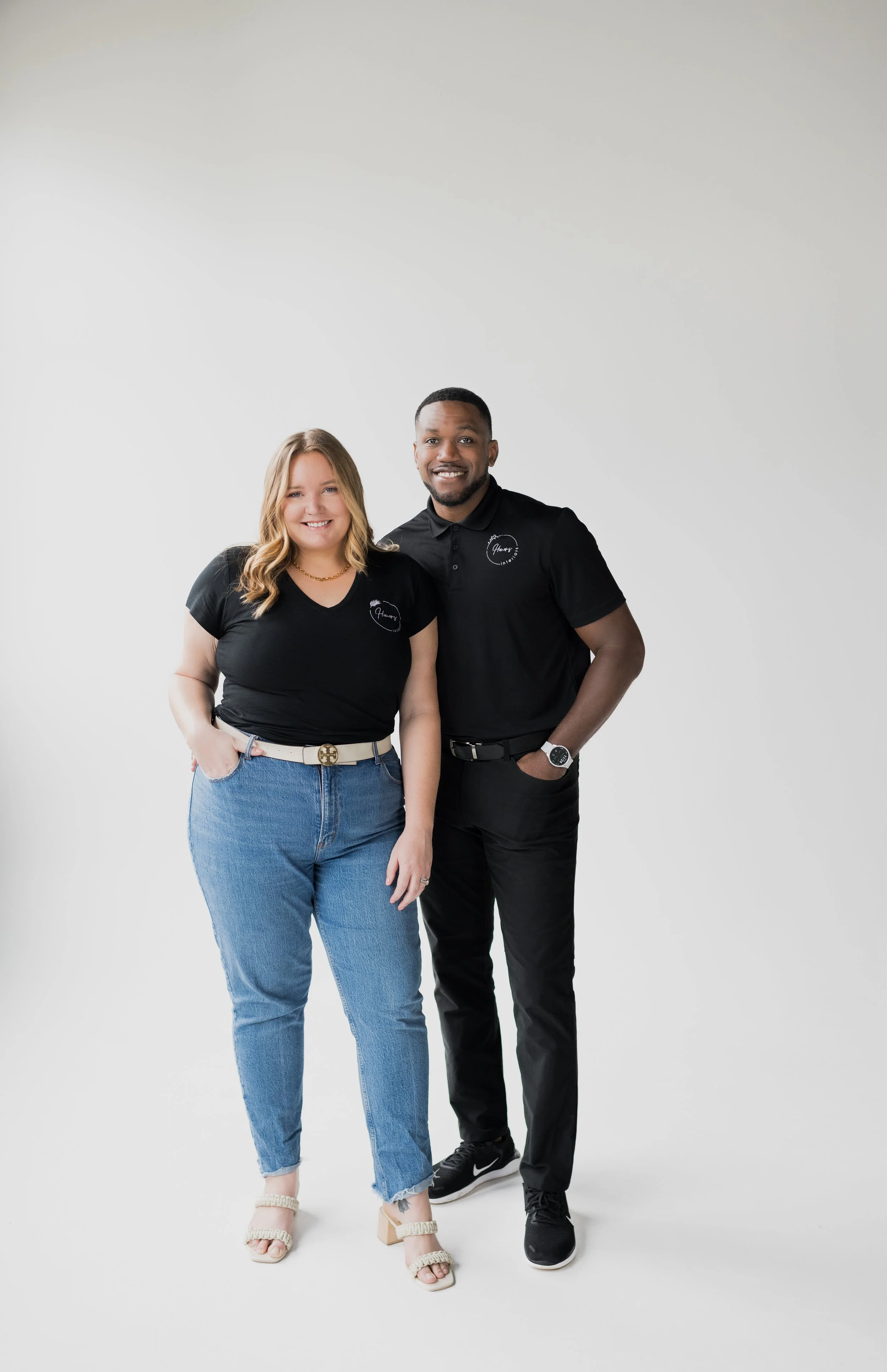 A smiling woman with blonde hair wearing a black T-shirt and blue jeans, standing next to a smiling man with short hair wearing a black polo shirt and black pants. They are standing against a plain white background and both are wearing casual shoes.