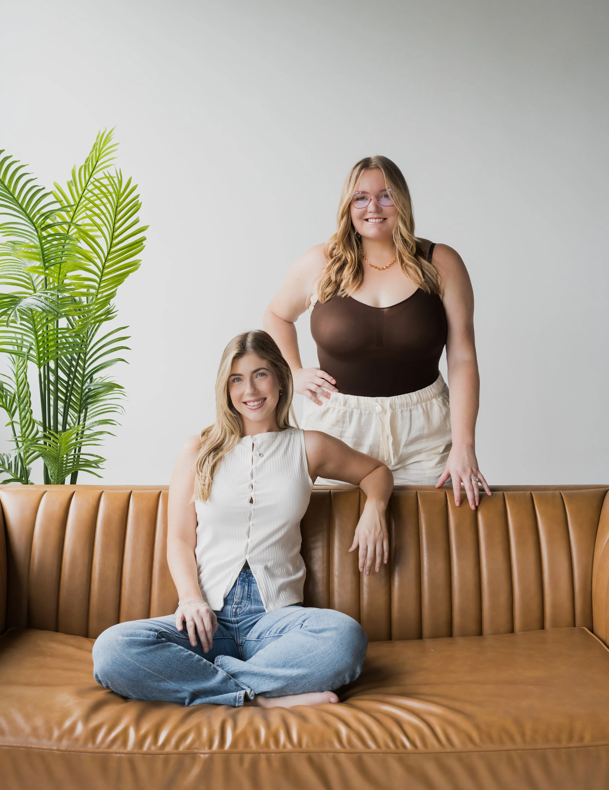 Two young women with light skin and blonde hair in casual clothing sitting and standing on a tan leather sofa in a bright room with a large green plant on the left side.