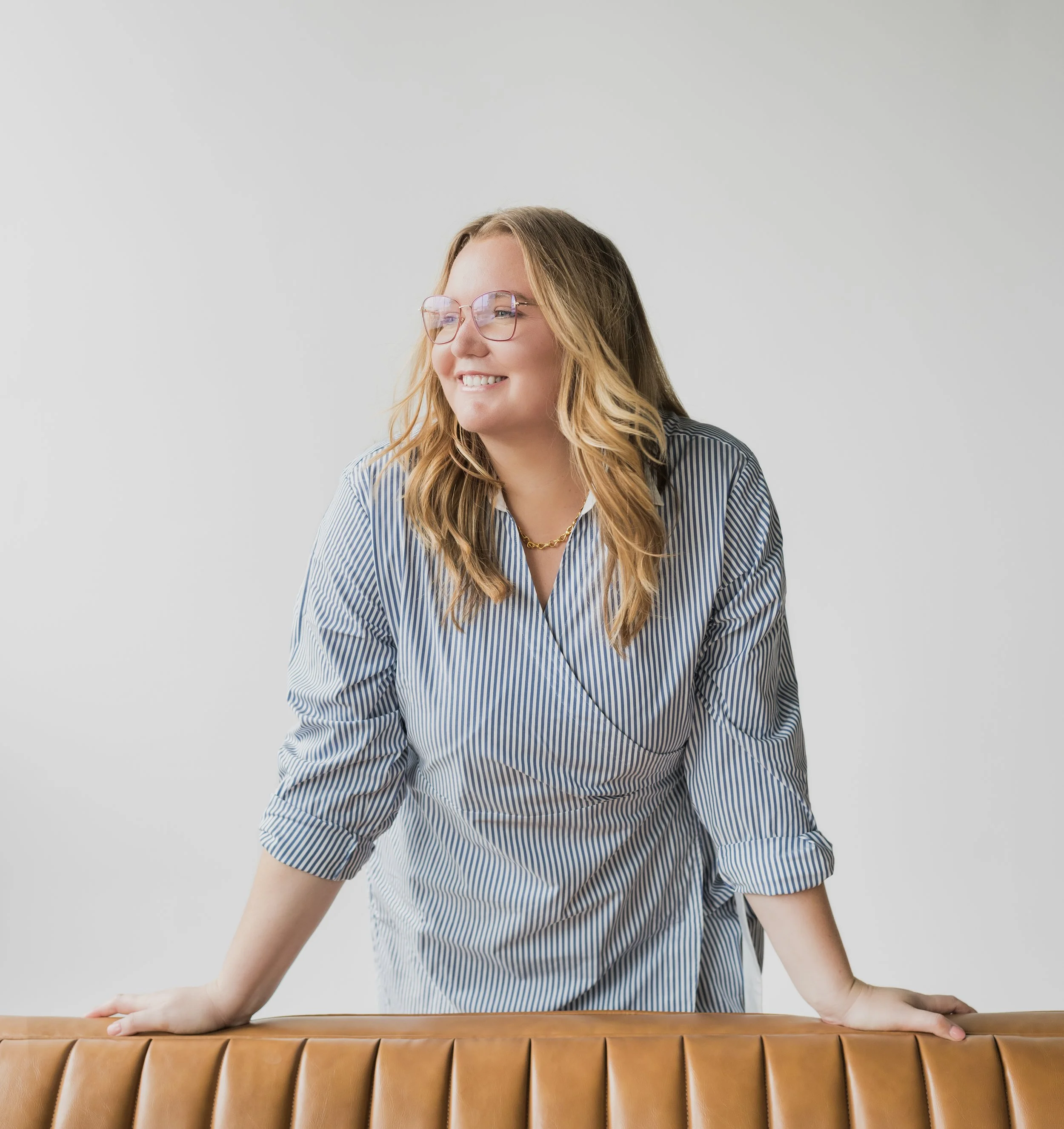 A woman with blonde hair wearing glasses and a blue striped blouse, leaning on a tan leather couch with a gray background.