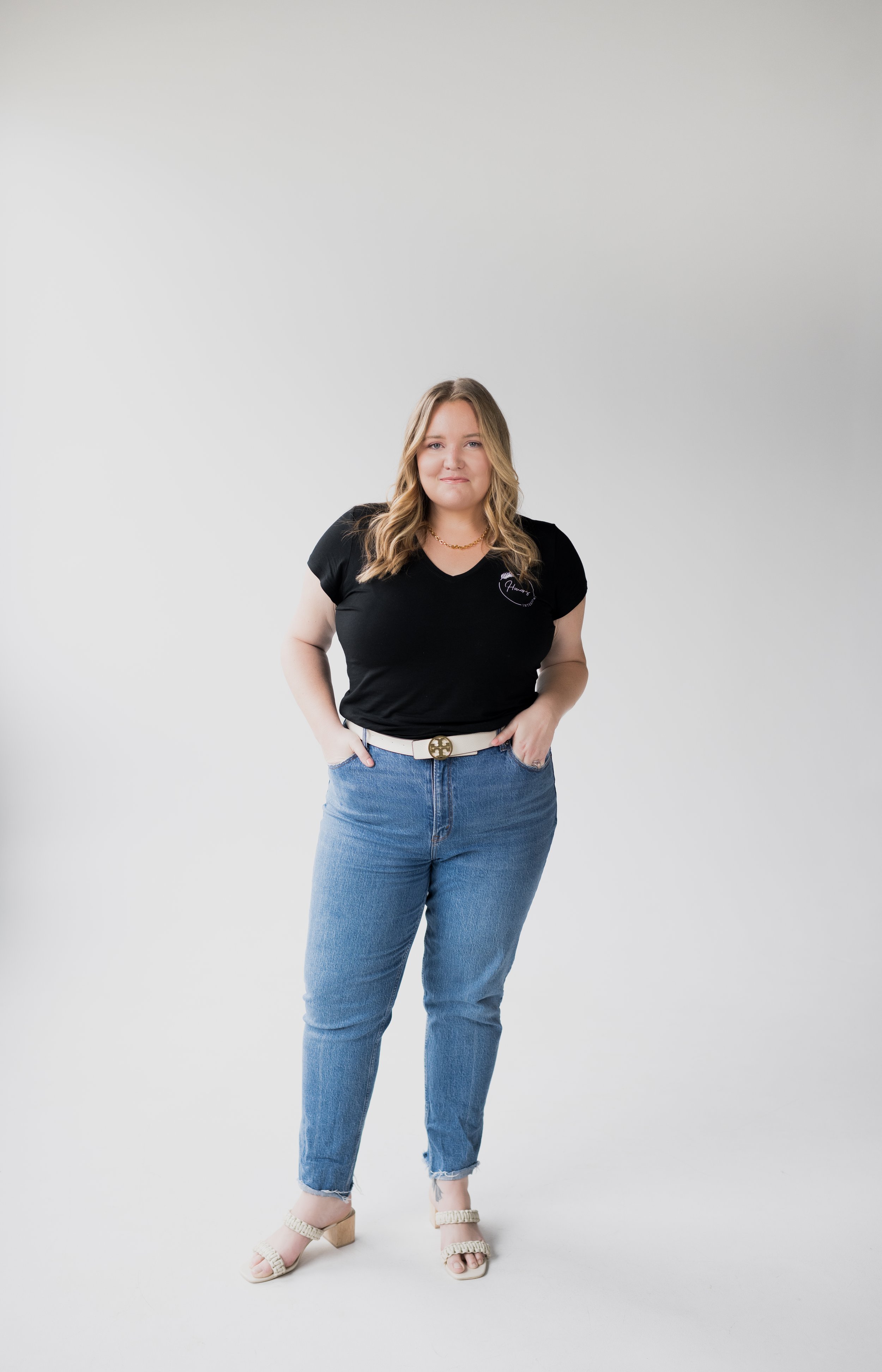 A woman with blonde hair wearing a black t-shirt, blue jeans, and beige heels posing against a plain white background.