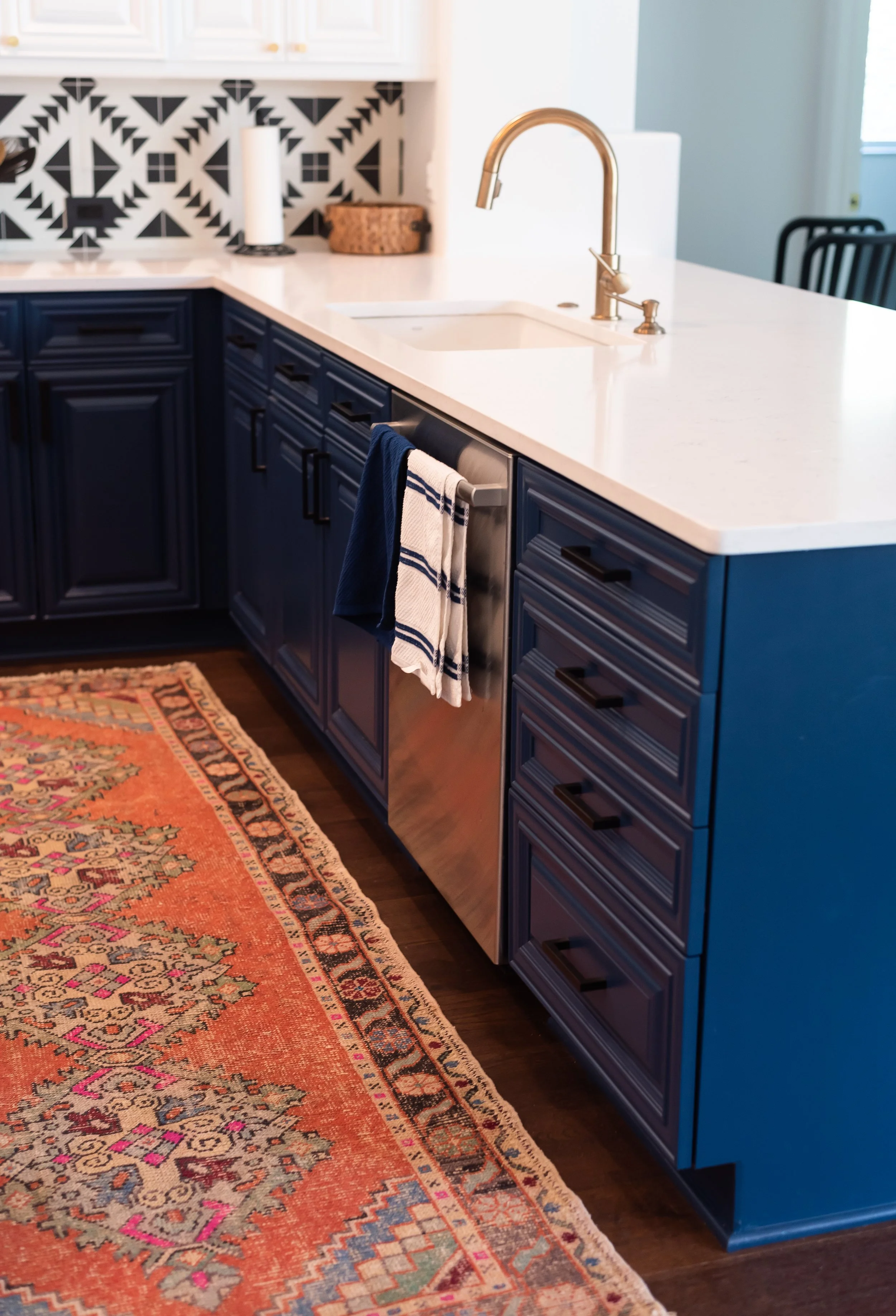 Kitchen with navy blue cabinets, white countertop, brass faucet, patterned tile backsplash, dish towels hanging, stainless steel dishwasher, white upper cabinets, colorful area rug, and black chairs in the background.