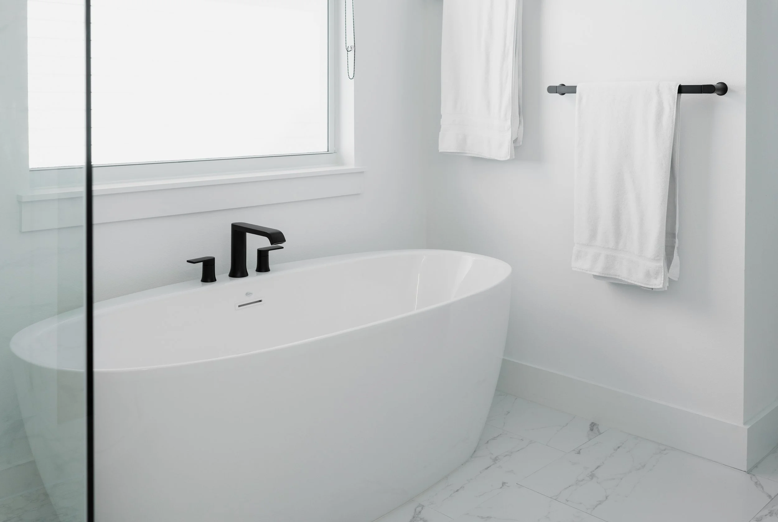 Modern bathroom with a white freestanding bathtub with black fixtures near a large window, white walls, and white towels hanging on a black towel rack.