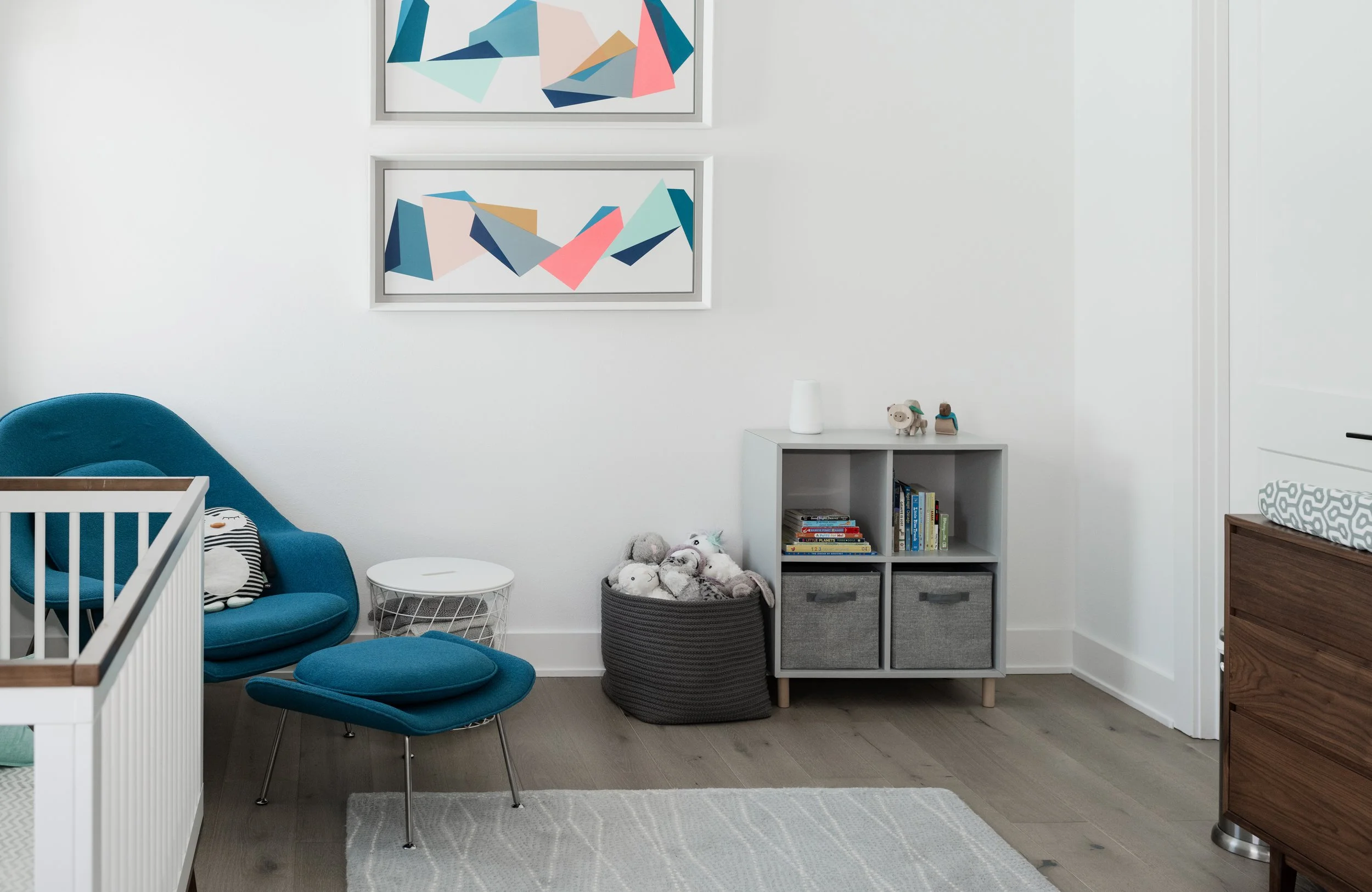 A cozy corner of a nursery with white walls, a blue chair and matching ottoman, a white side table, a gray storage cube filled with plush toys, a small bookshelf with children's books, and abstract framed artwork on the wall.