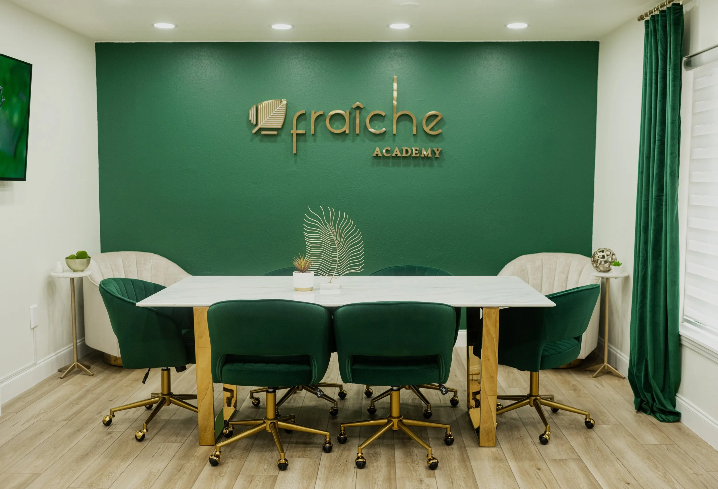 Meeting room with green accent wall, gold lettering sign says 'fraiche ACADEMY', white marble table, six green swivel chairs, beige side chairs, and green curtains.