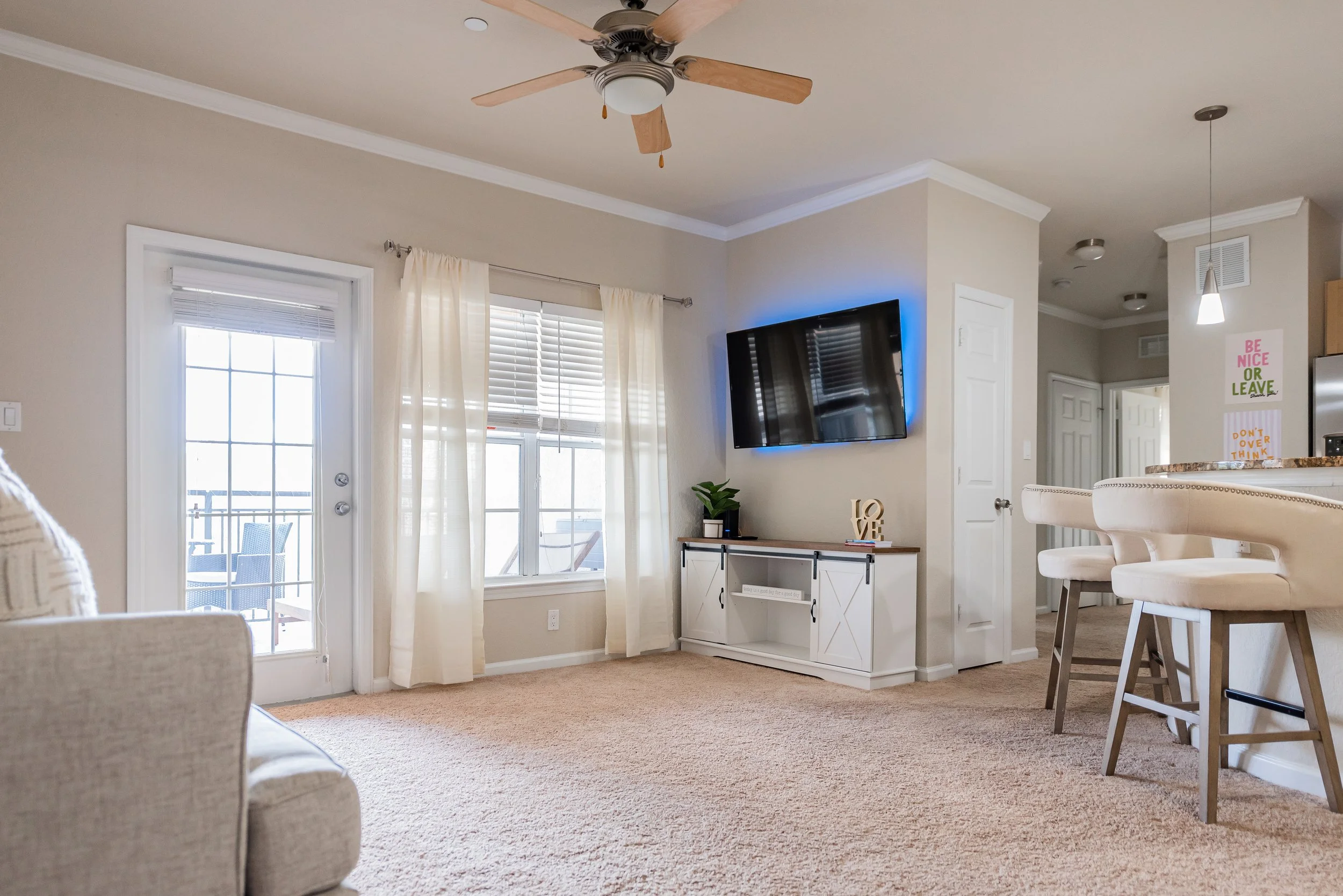 Living room with beige walls, a ceiling fan, a flat-screen TV mounted on the wall, a white TV stand, a plant, two chairs in the kitchen area, and a glass door leading to a balcony with patio furniture.