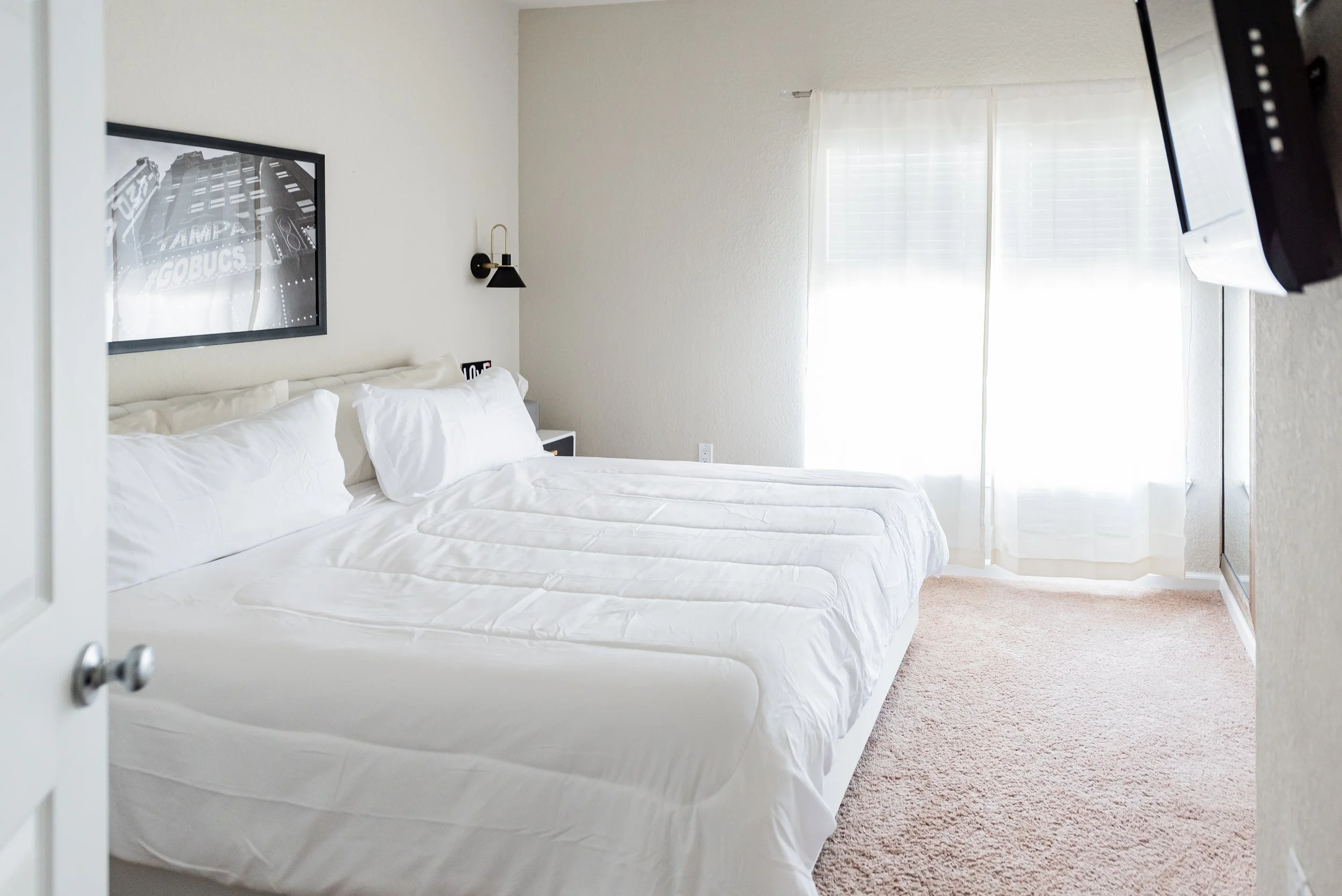Clean, white bedroom with a king-sized bed, a black and white framed picture of a building above the bed, black wall-mounted light fixture, and large windows with white curtains letting in natural light.