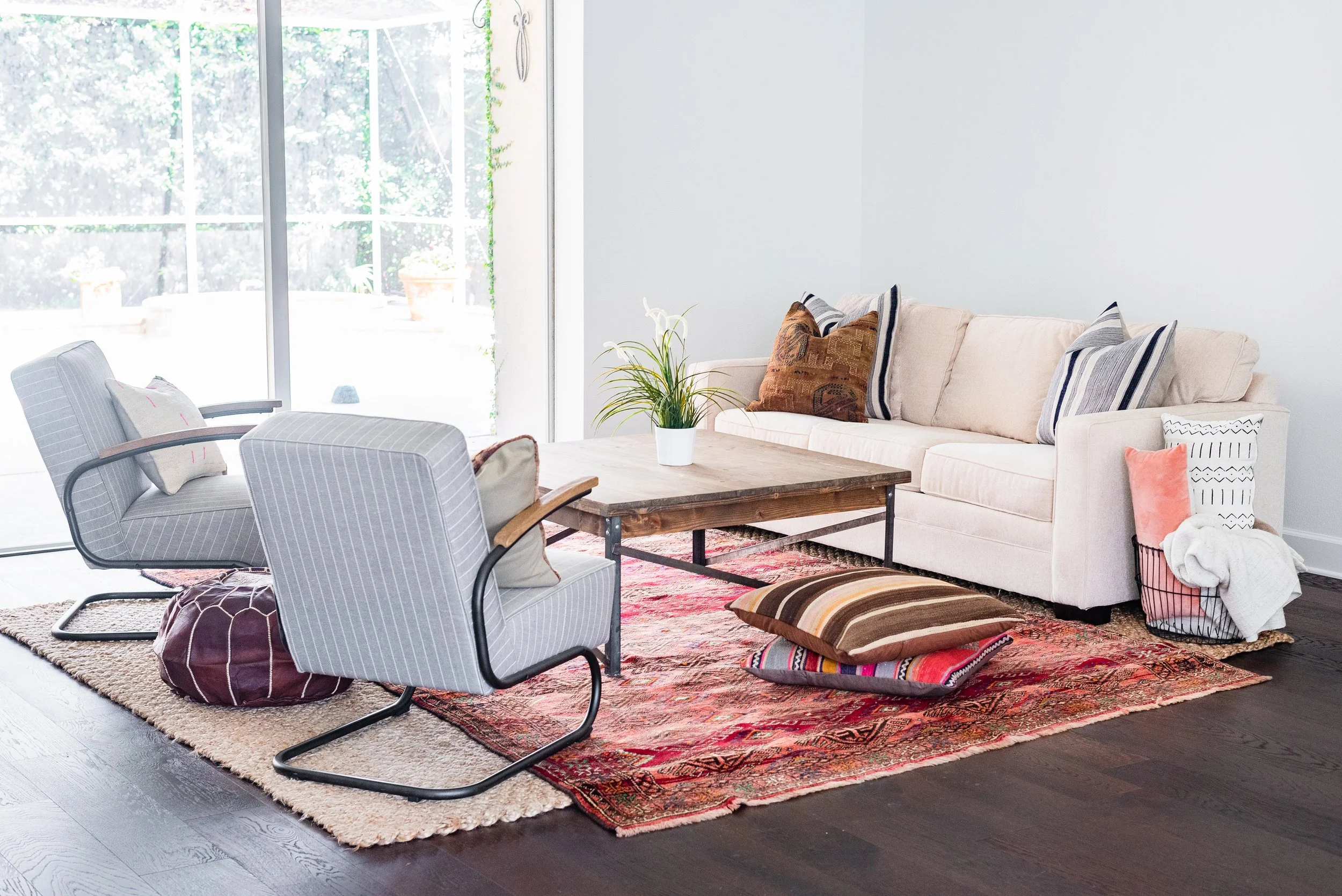A living room with a white sofa, two striped armchairs, a wooden coffee table, and various patterned cushions. There's a plant on the coffee table, a recessed area with baskets, and a large sliding glass door letting in natural light.