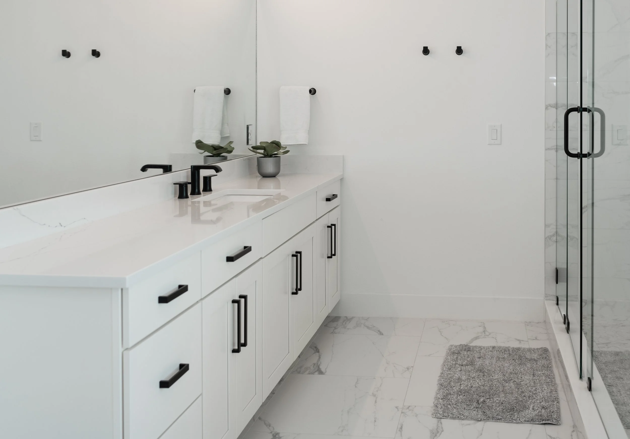 Modern bathroom with white vanity, black fixtures, large mirror, potted plant, white towels, marble floor, glass shower door, and gray bath mat.