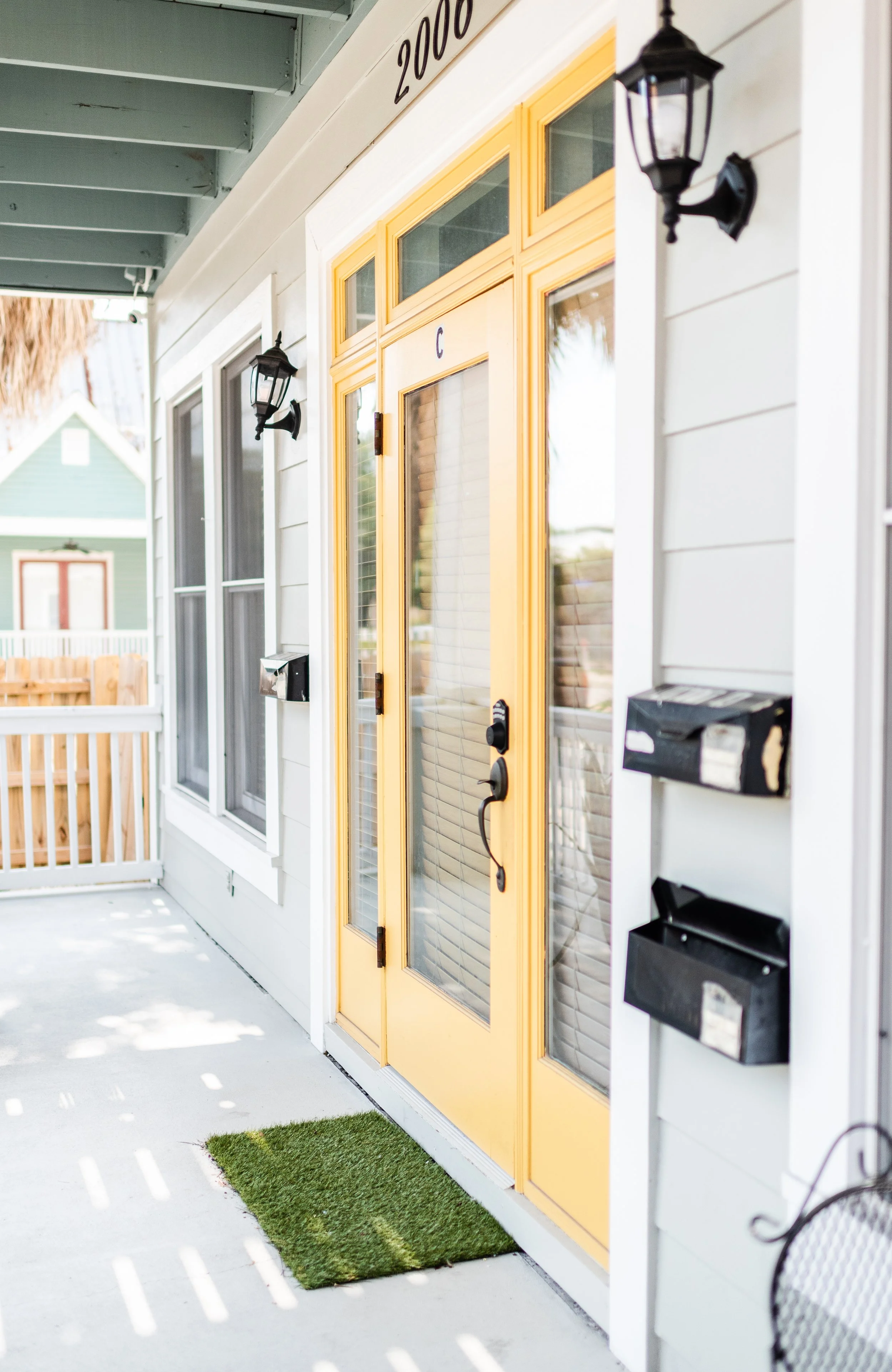Front porch of a house with a yellow door, outdoor black lanterns, mailboxes, and house number 2000, with neighboring houses in the background.