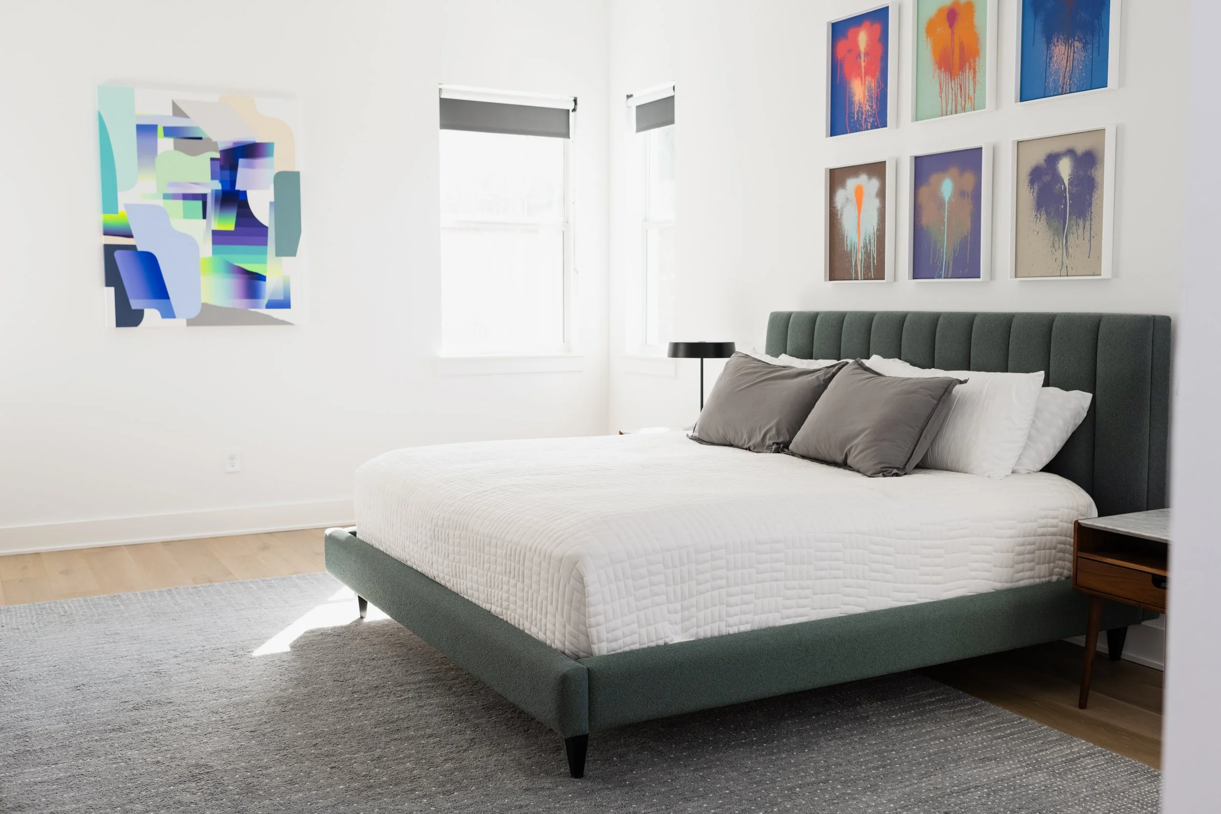 A modern bedroom with a green upholstered bed, white bedding, gray and white pillows, artwork on the white walls, two windows with black blinds, a black bedside lamp, and a gray rug on wooden flooring.