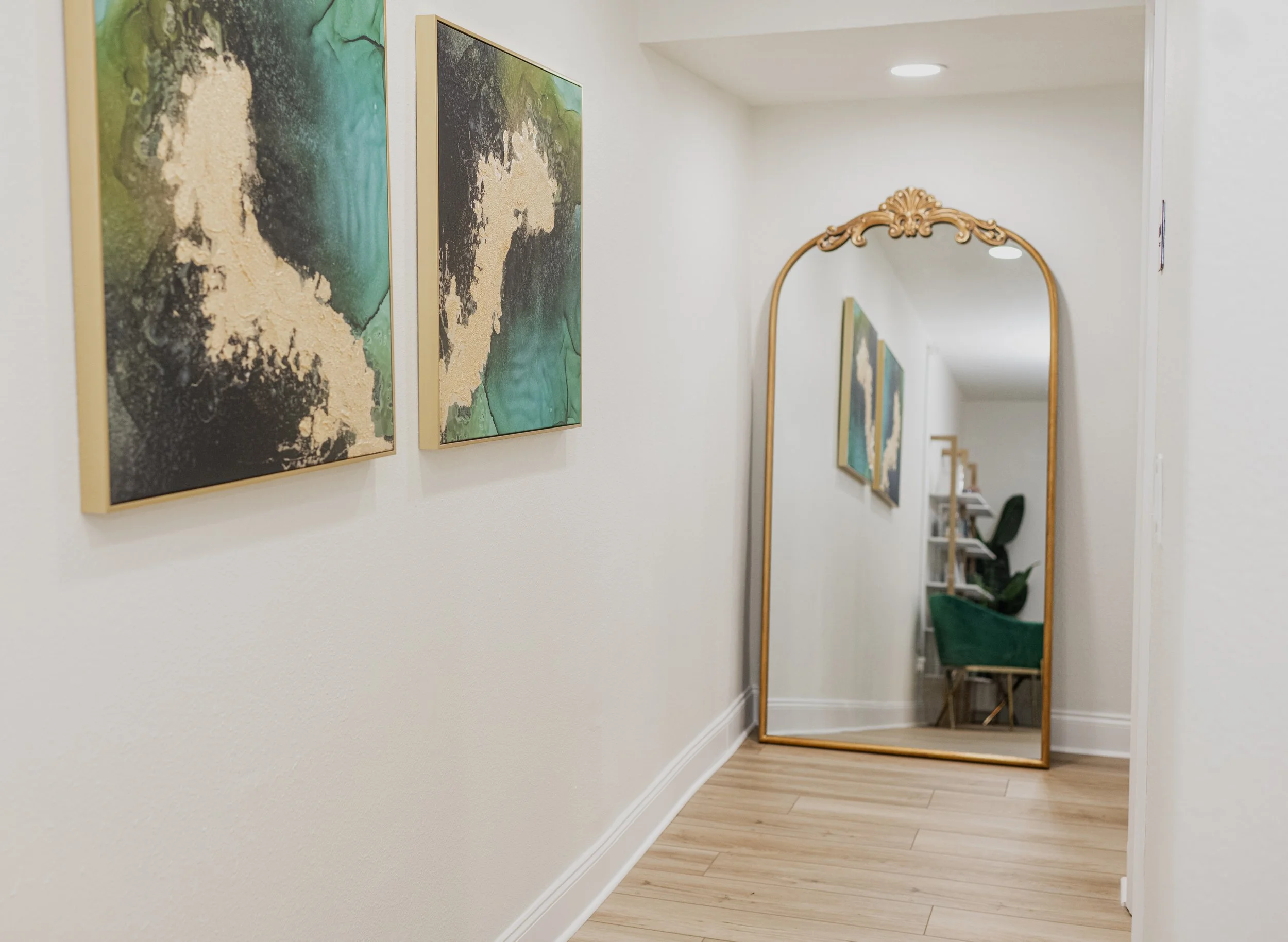 A hallway with light-colored walls and wooden flooring featuring two framed abstract paintings on the left wall, a large decorative gold-framed mirror on the right, reflecting chairs and a bookshelf in the background.