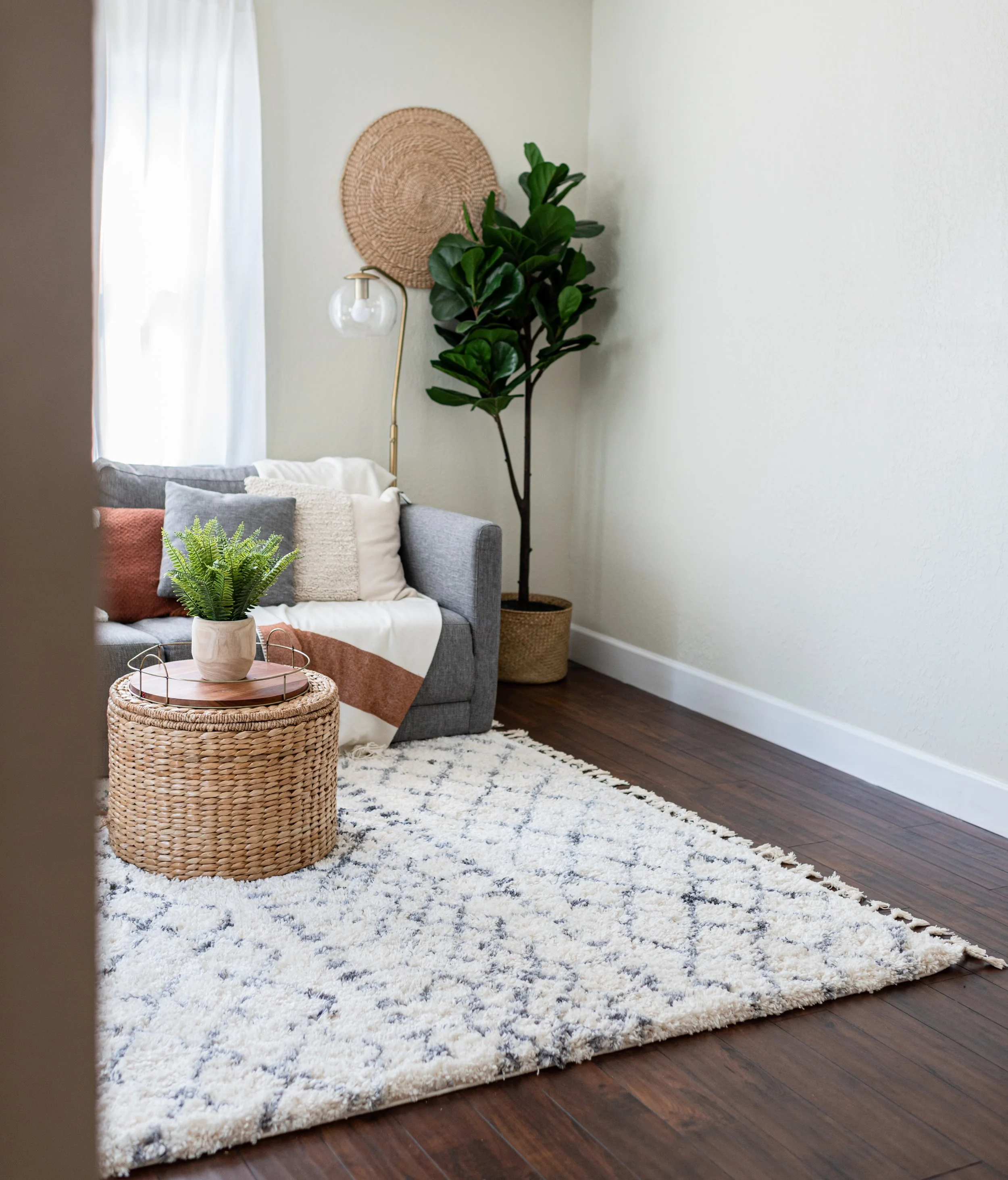 Living room with a gray sofa adorned with throw pillows, a textured white and rust blanket, a round wicker ottoman with a potted fern on top, a cream and navy rug, and a large green plant in a woven basket. Light filters through cream curtains.