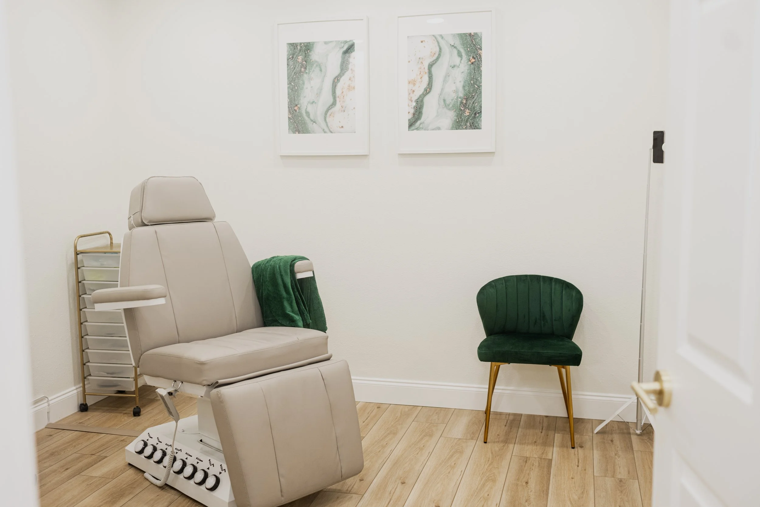 A medical examination room with a beige reclining chair, a small green velvet chair, two framed abstract art pieces on the white wall, a small rolling storage cart, and a white door partially open.