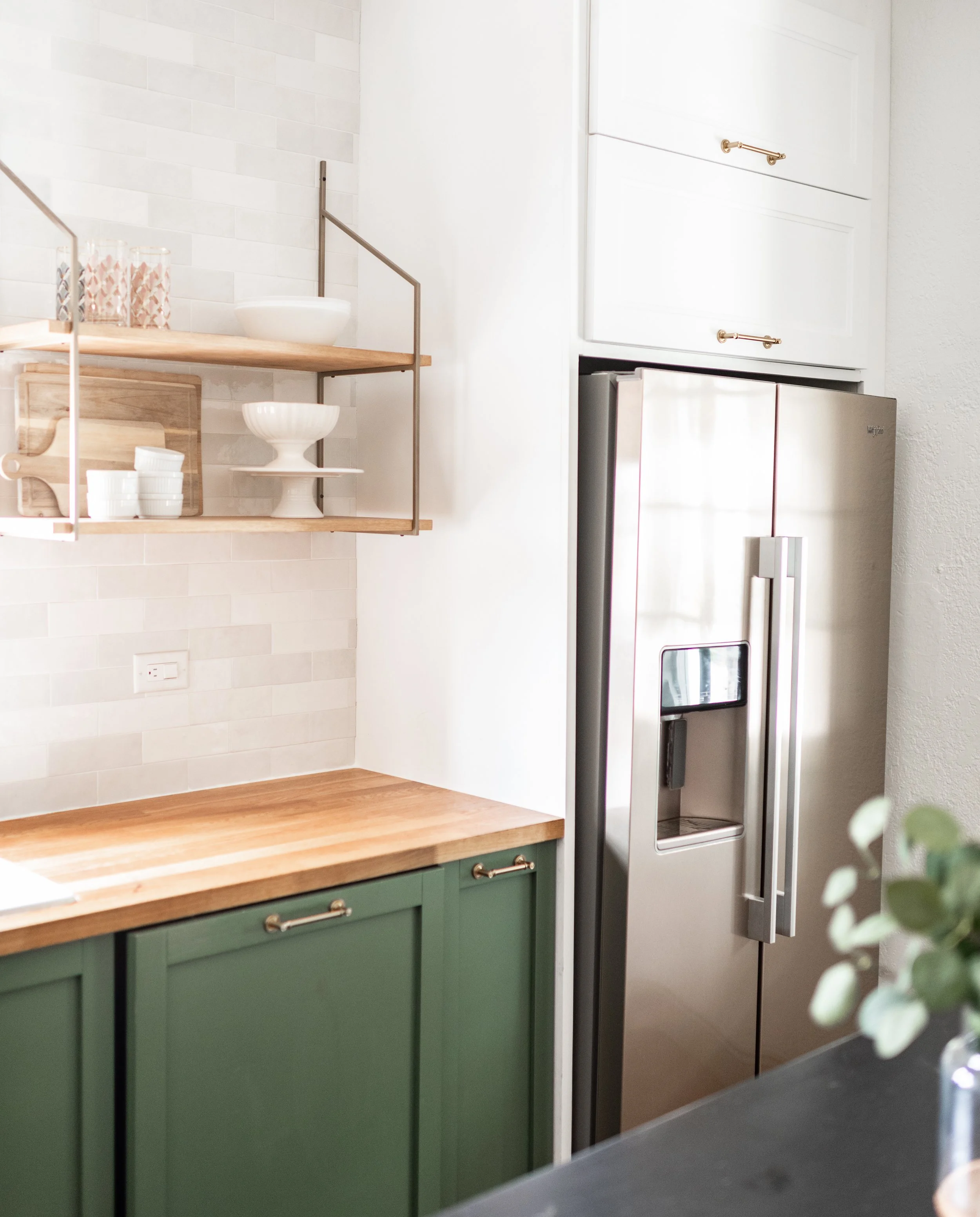 Kitchen with green cabinets, wooden countertop, stainless steel refrigerator, open shelving with dishes, white wall tiles.