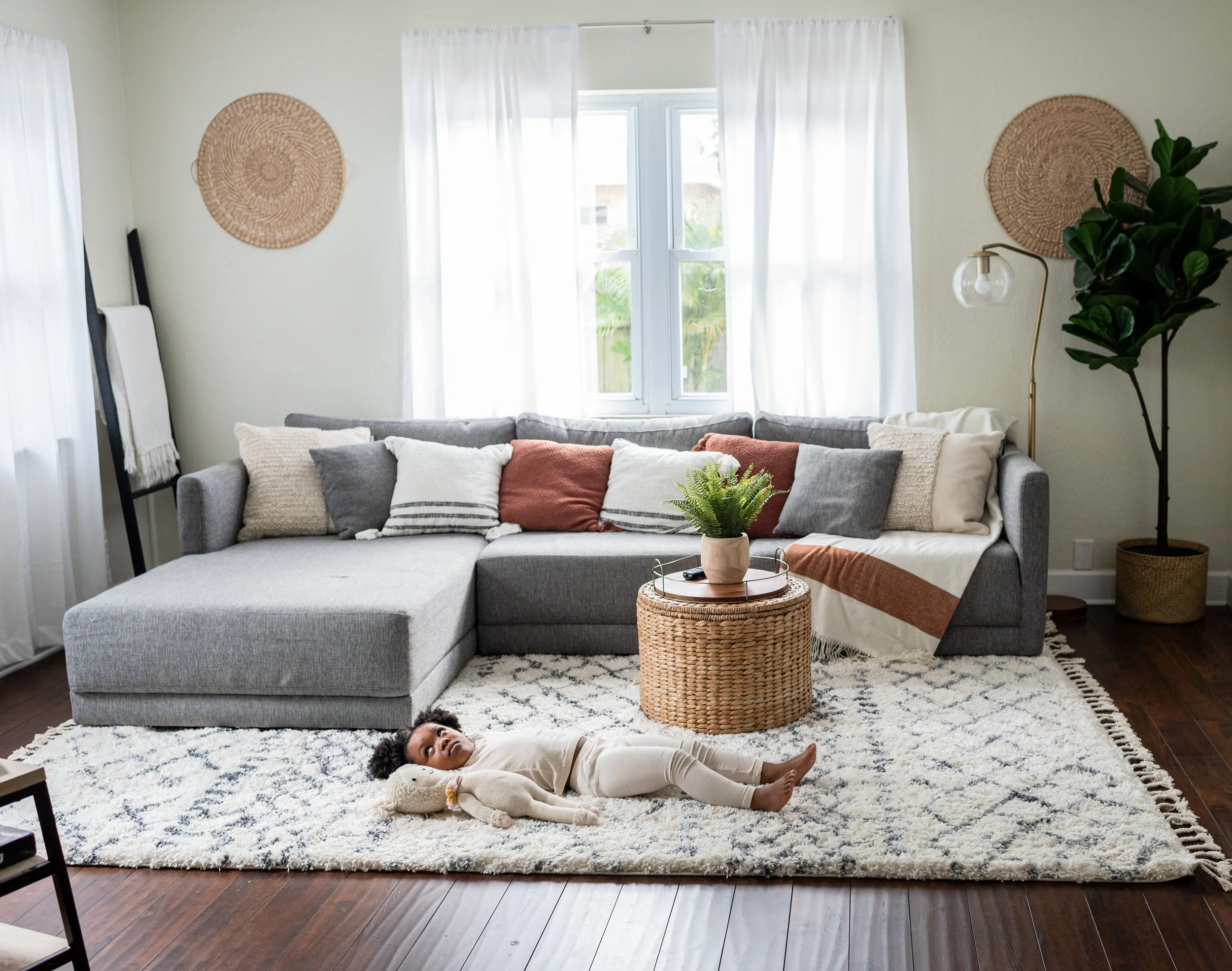 A living room with a large gray sectional sofa, a round wicker coffee table with a plant, and pillows. A young girl lies on a white and navy rug, playing with a stuffed rabbit. There is a window with white curtains, a tall green plant in a woven bask