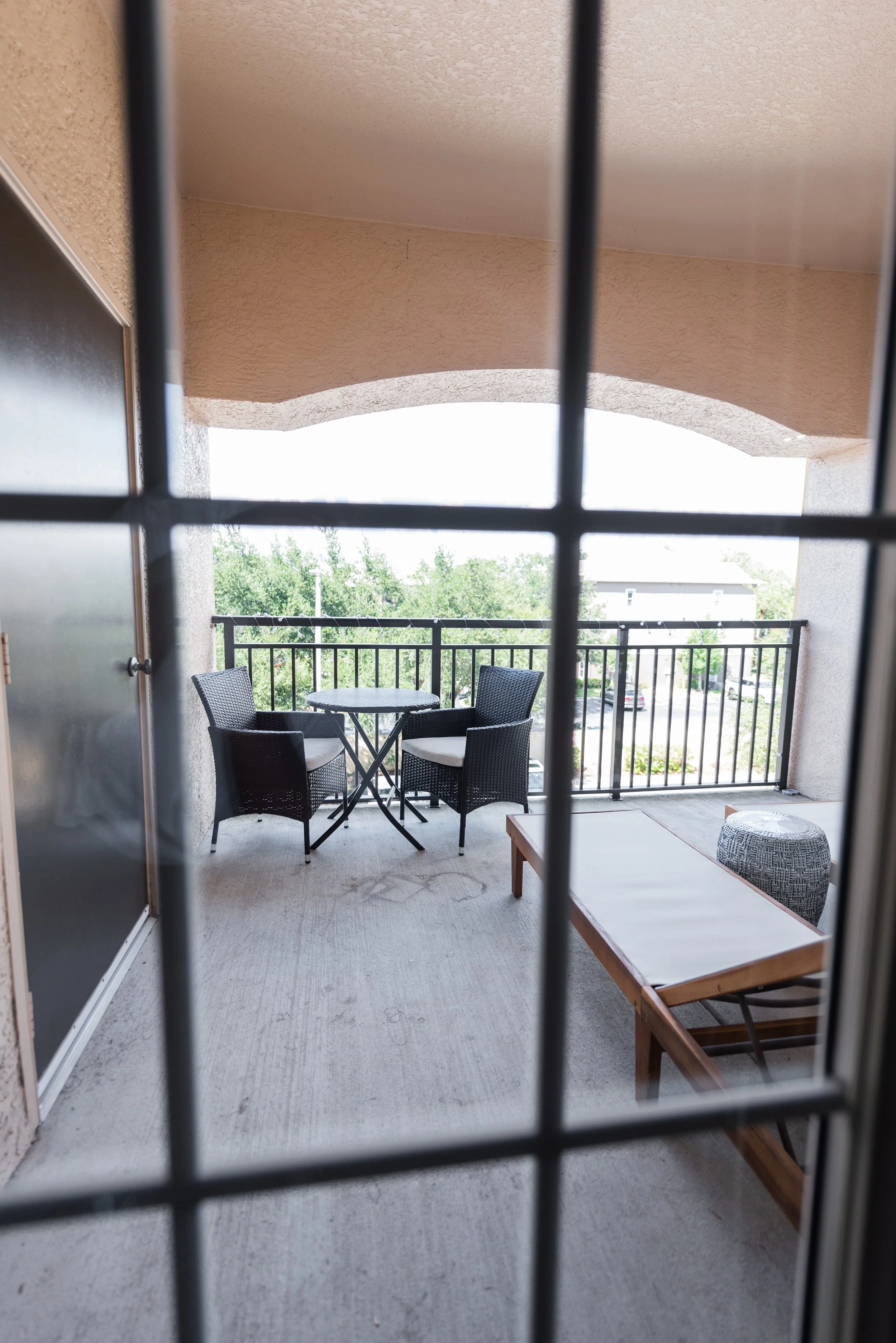 View of a balcony seen through a window with black grid pattern. The balcony has a small black round table with four black wicker chairs, a wooden lounge chair with a white cushion, and a gray ottoman. The background shows trees and a parking lot wit