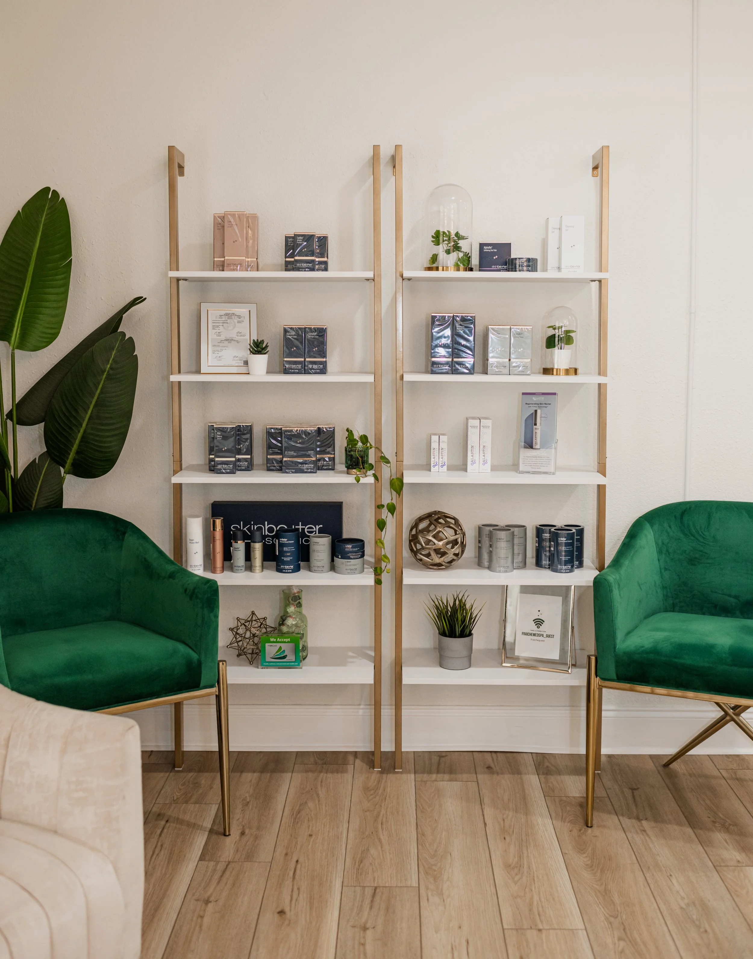 Empty shelving units with skincare products, decorative items, and plants, flanked by two green velvet chairs in a modern waiting area.
