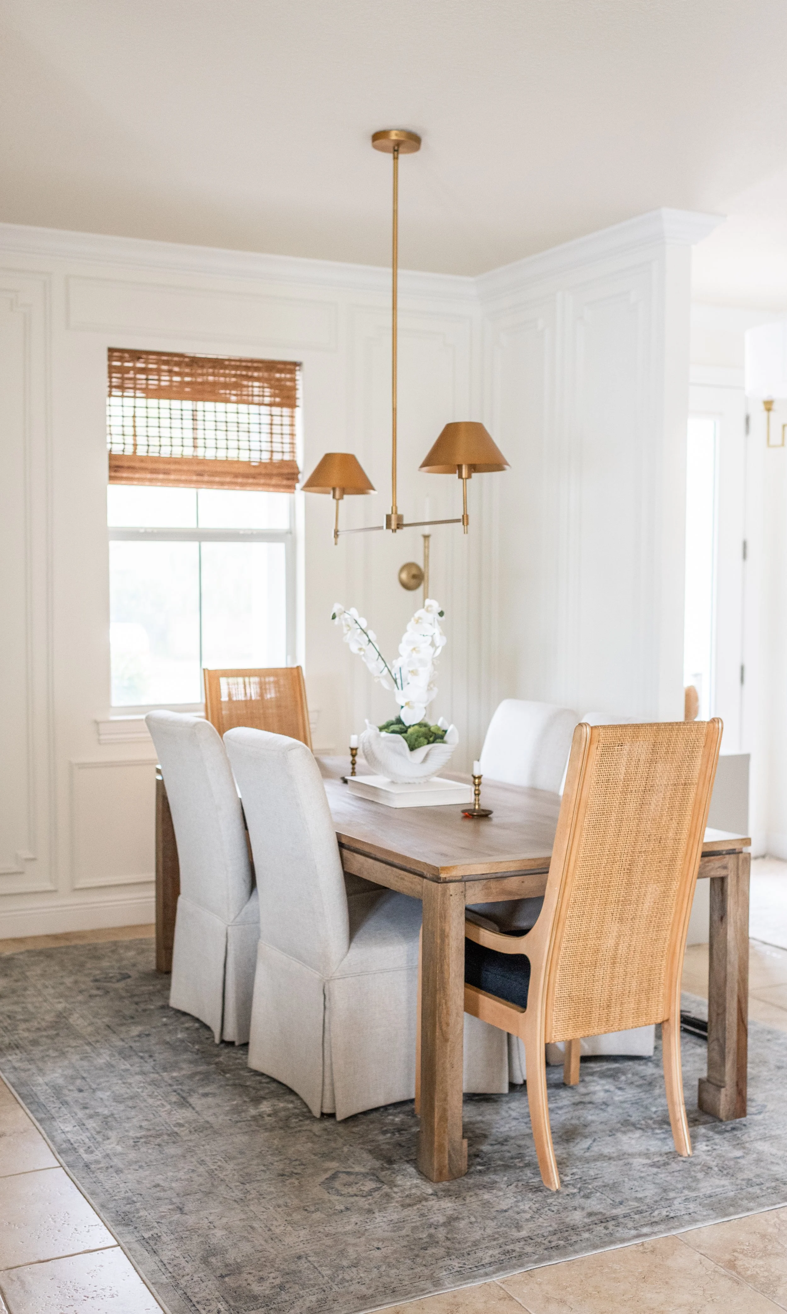 A dining room with a wooden table, six chairs—two white upholstered and four rattan-backed. There's a centerpiece with white orchids on the table, a window with a bamboo shade, a patterned area rug, and a hanging brass and wood chandelier.